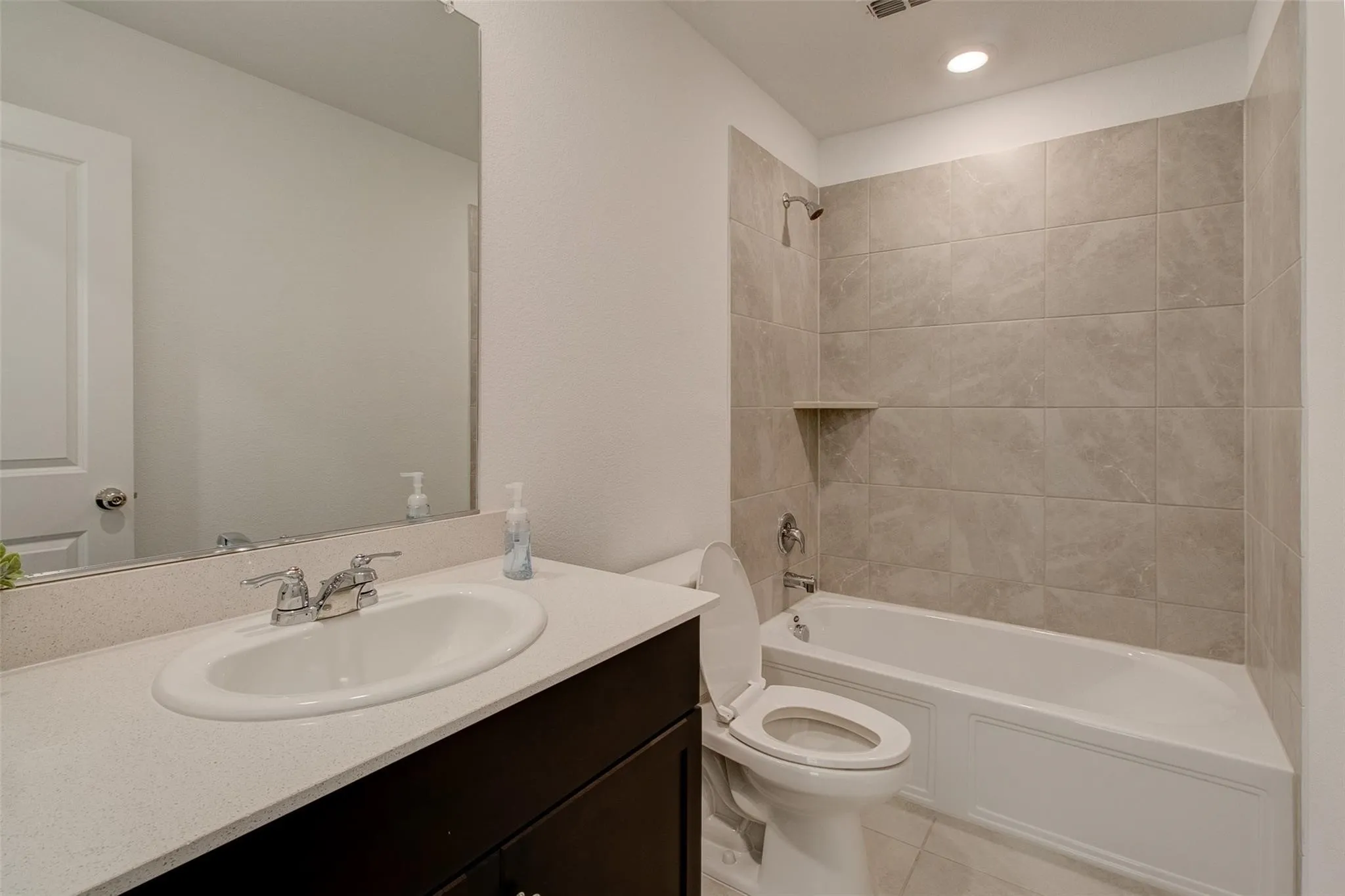 Bathroom featuring a single vanity with a white countertop and dark wood cabinetry, an undermount sink with chrome fixtures, a full-width mirror, and a bathtub with light gray tiled surround and built-in​​‌​​​​‌​​‌‌​‌‌​​​‌‌​‌​‌​‌​​​‌​​ shelf