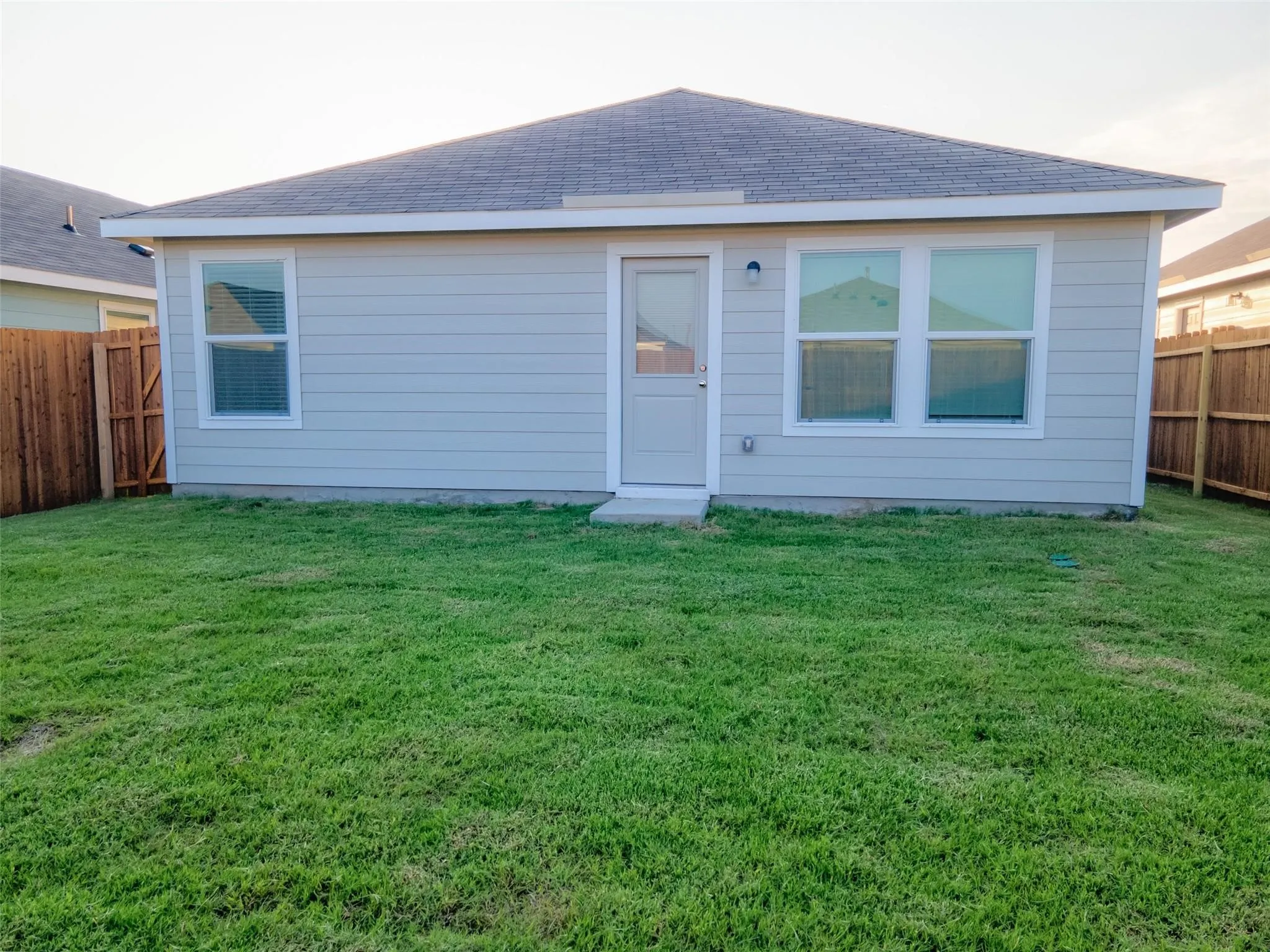 Spacious backyard with a vibrant green lawn, light-colored siding, multiple windows, a rear entry door, and perimeter fencing