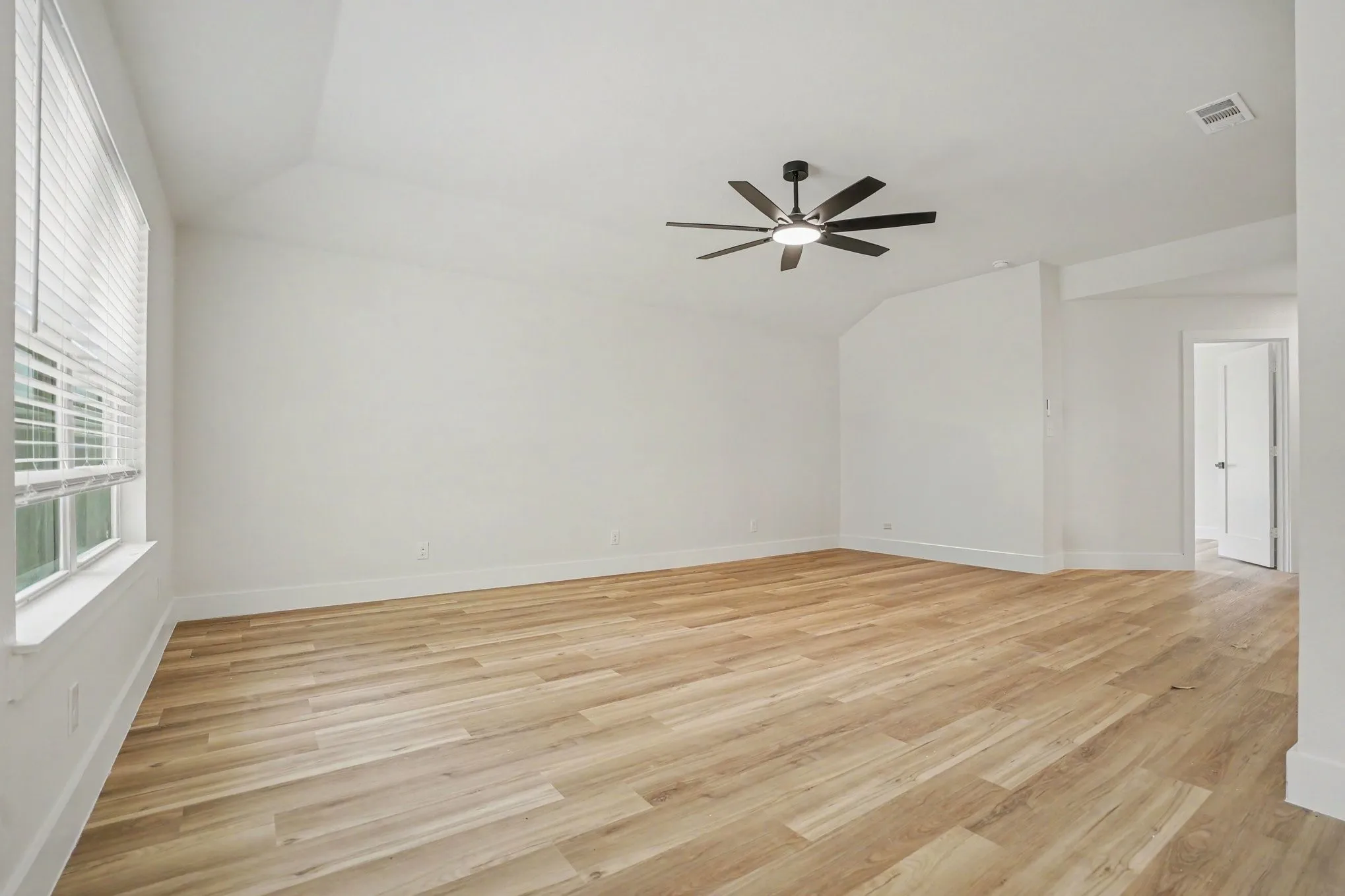 Spacious bedroom with warm wood-finish flooring throughout, abundant natural light from large windows with plantation shutters, and a modern ceiling fan with integrated lighting fixture
