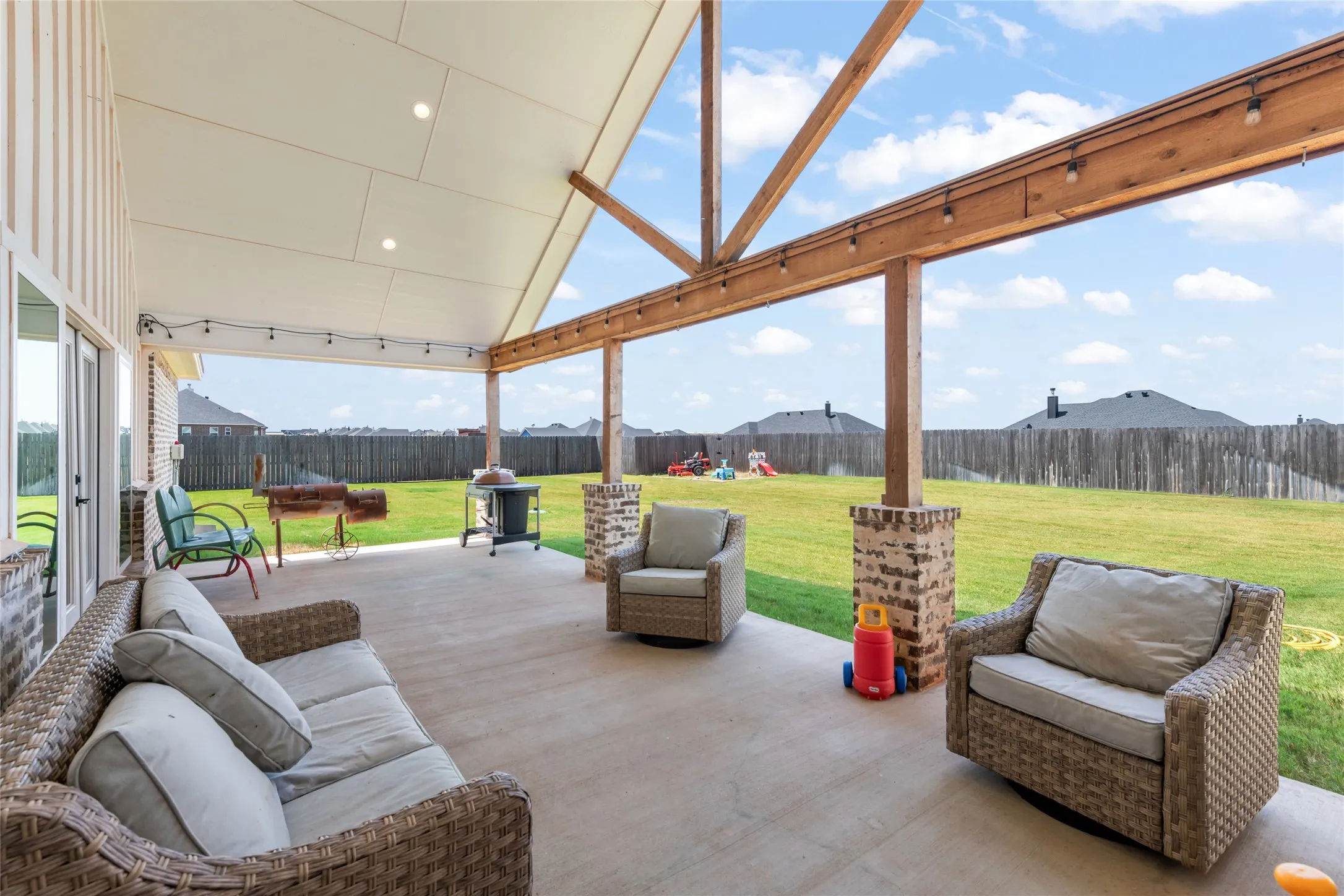 Expansive covered patio featuring a vaulted ceiling with recessed lighting, exposed wood beams, brick-clad support columns, and string lighting