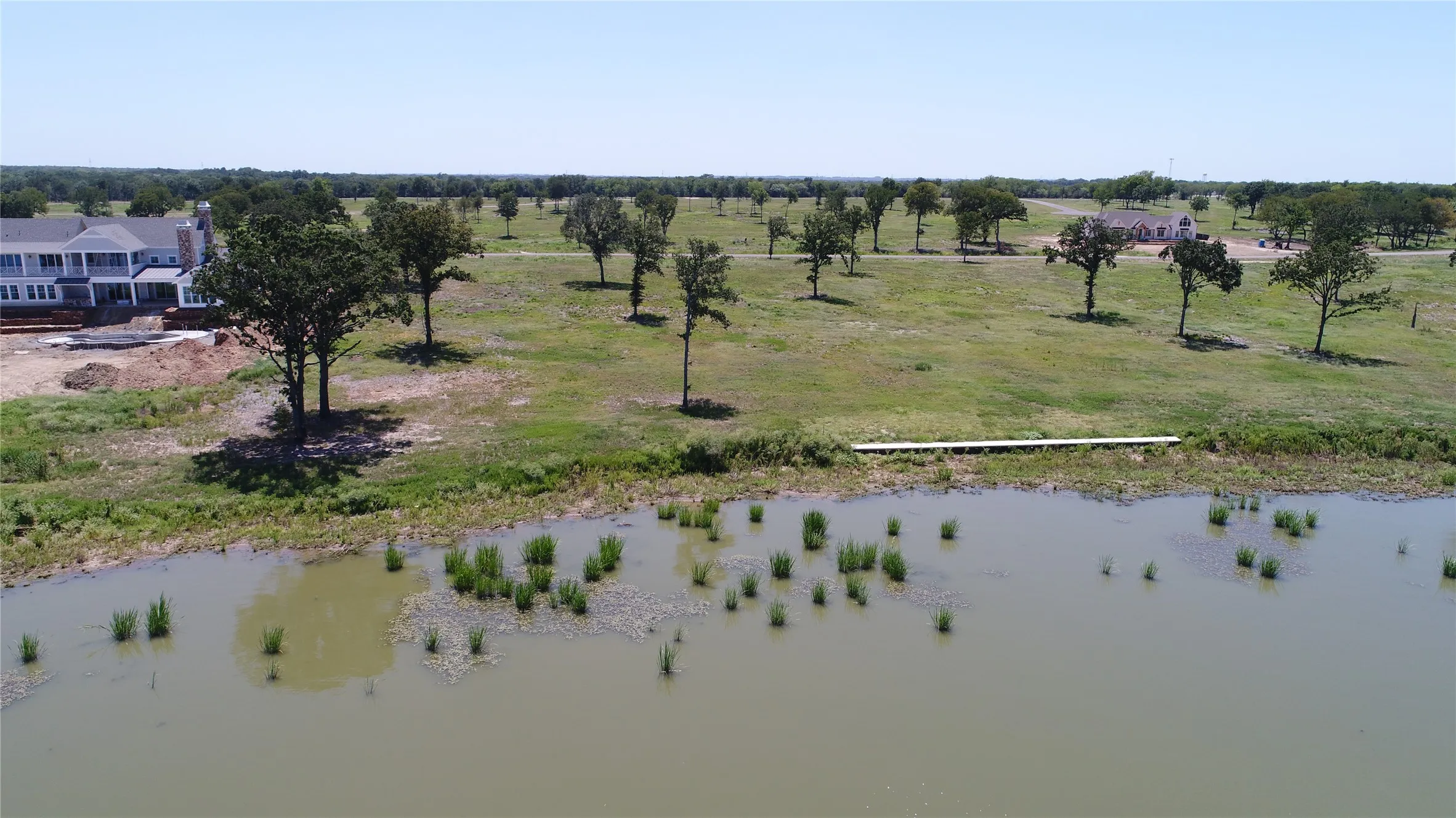 Expansive lakeside property featuring a private dock, mature trees, and a distant residence under construction