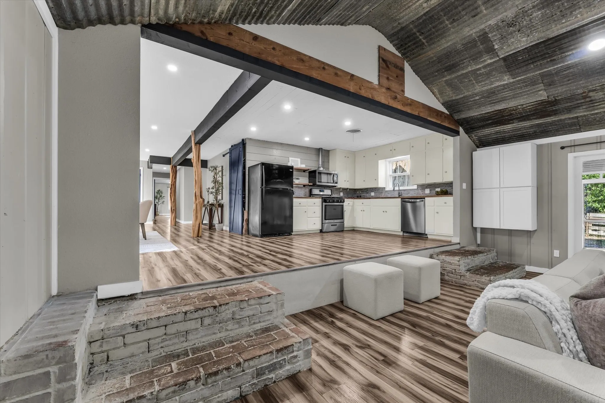 Kitchen and living area featuring exposed wood beams, corrugated metal ceiling, wood-finish flooring, white cabinetry, and stainless steel​​‌​​​​‌​​‌‌​‌‌​​​‌‌​‌​‌​‌​​​‌​​ appliances