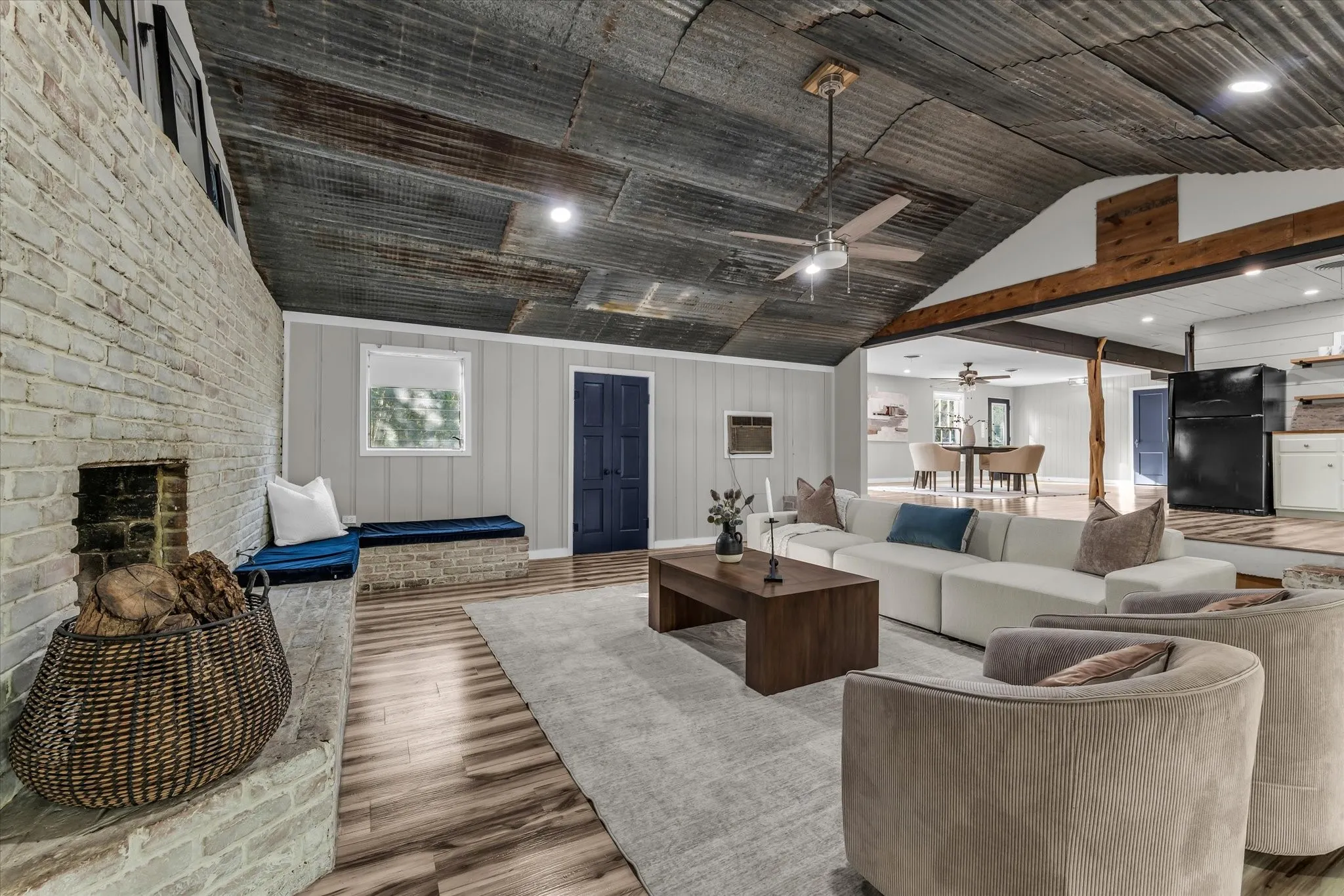 Living room featuring a corrugated metal vaulted ceiling, wood-finish flooring, and a whitewashed brick fireplace