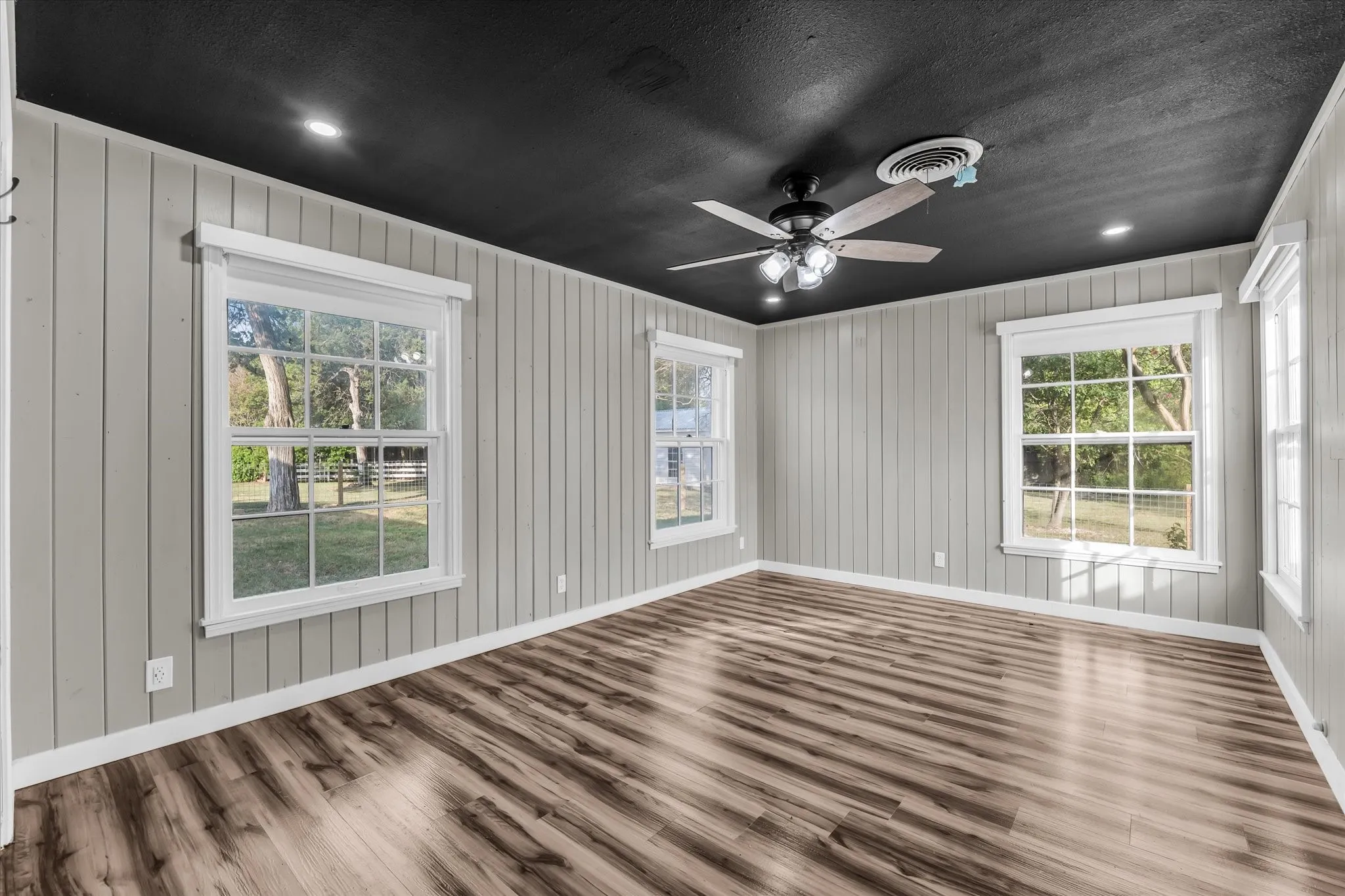 Spacious interior room featuring wood-finish flooring, vertical paneling walls, multiple windows with white trim, a dark ceiling, and a ceiling fan with integrated lighting