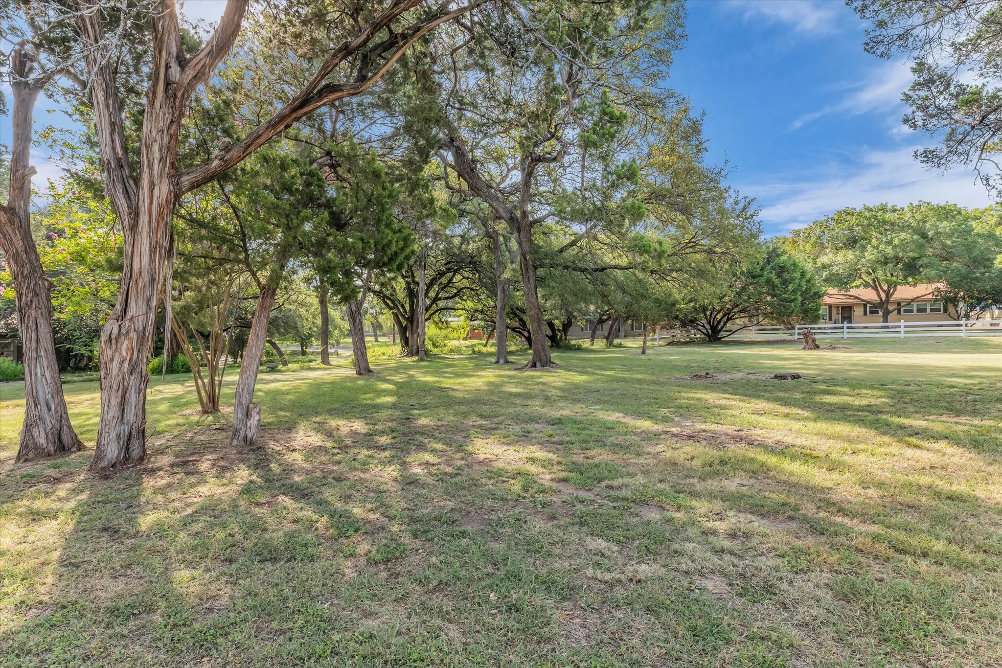 Expansive grassy area with numerous mature trees, a white picket fence, and a single-story residence in the background