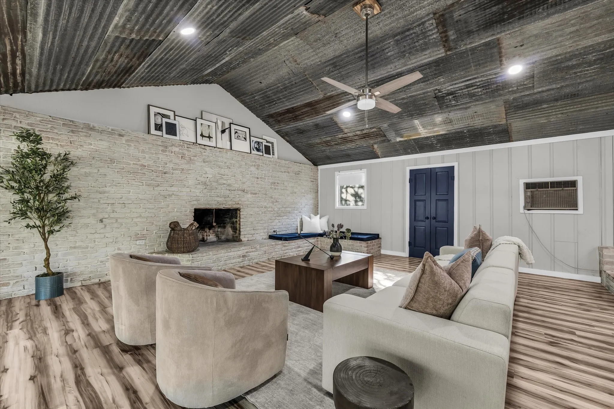 Living area featuring a corrugated metal ceiling, wood-finish flooring, a white brick fireplace, and a ceiling fan with integrated lighting