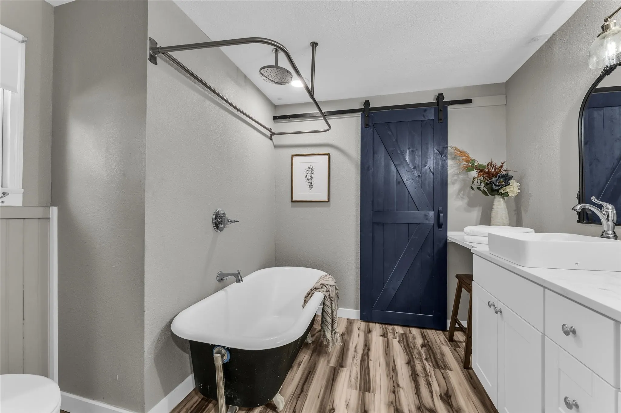 Vintage-style clawfoot tub with an overhead shower, wood-finish flooring, a contemporary vanity with a vessel sink, a dark blue sliding barn door, and beadboard paneling