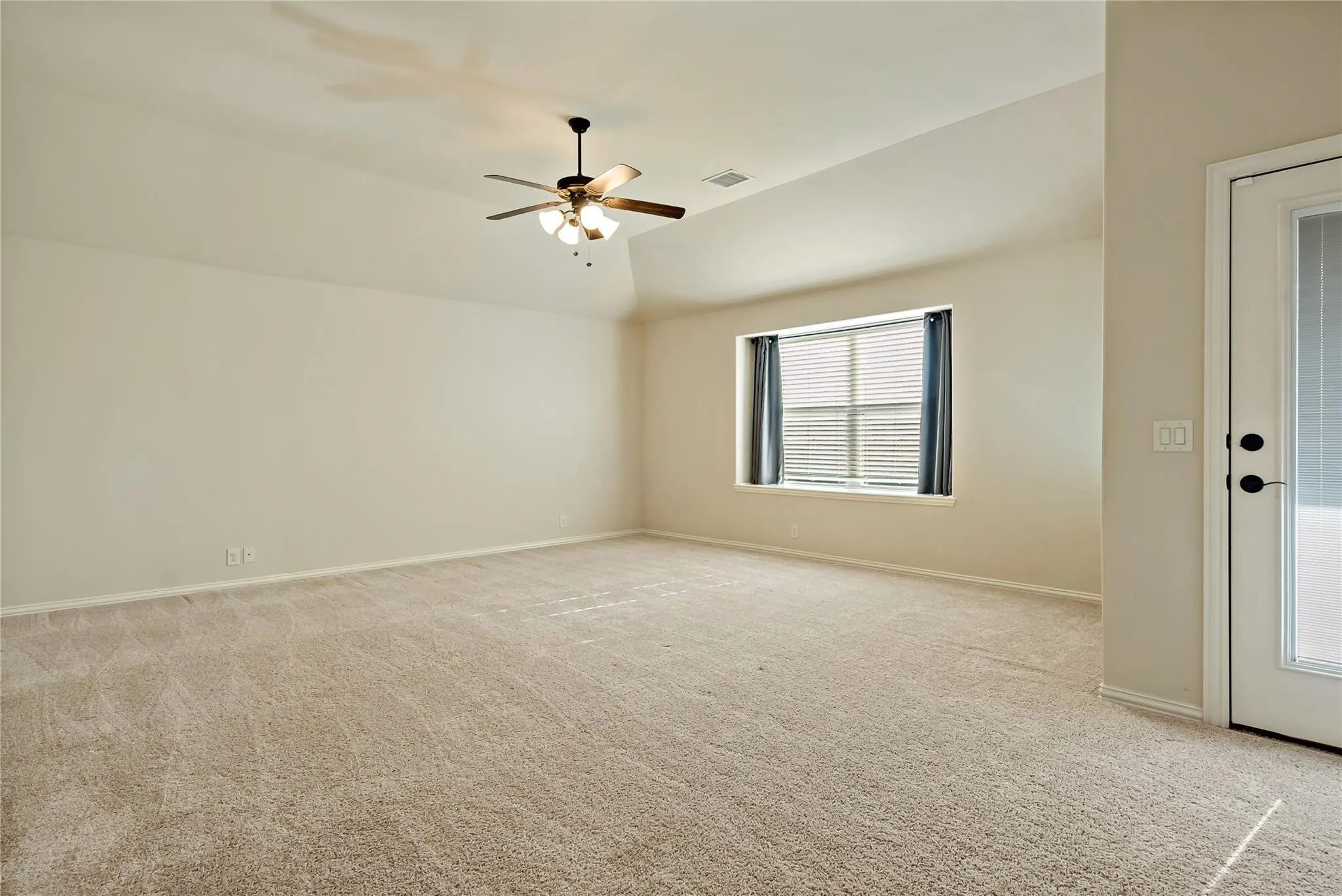 Spacious room featuring vaulted ceilings, neutral-toned carpeting, a ceiling fan with integrated lighting, and a large window with blinds and drapery