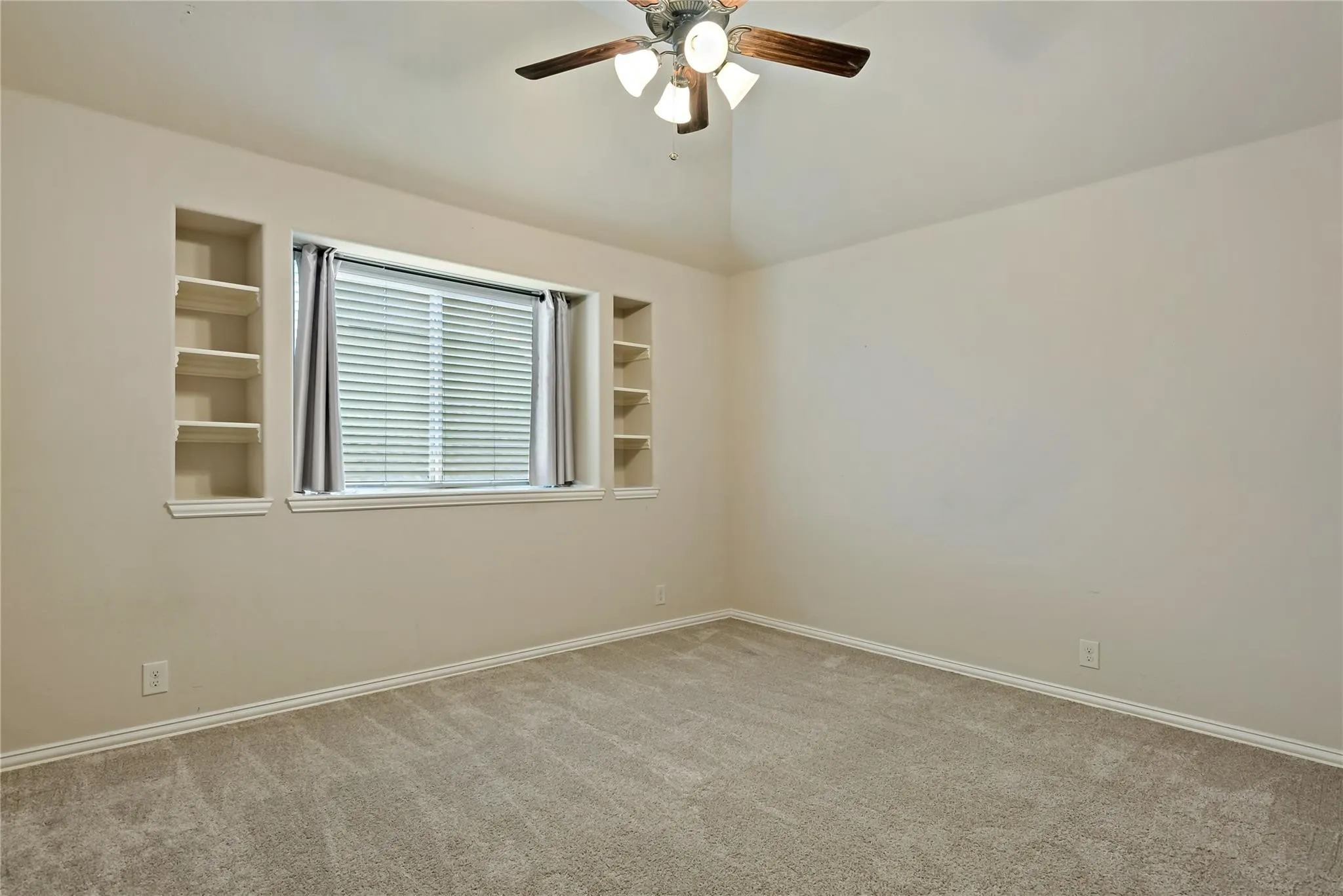 Carpeted room featuring a window with blinds, two built-in shelving units, a ceiling fan with integrated lighting, and neutral wall tones
