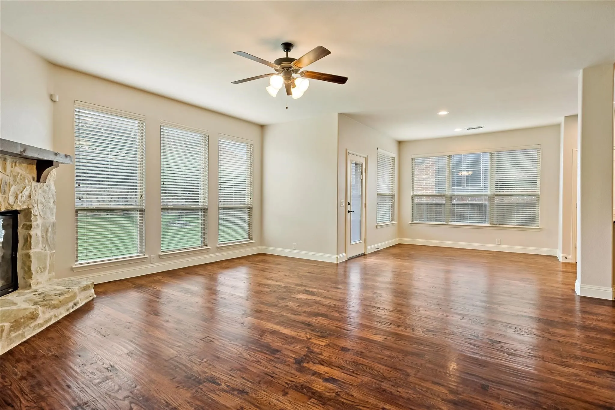 Spacious living area showcasing gleaming wood-finish flooring, abundant natural light from multiple multi-pane windows, a ceiling fan with integrated lighting, and a stone fireplace accent wall