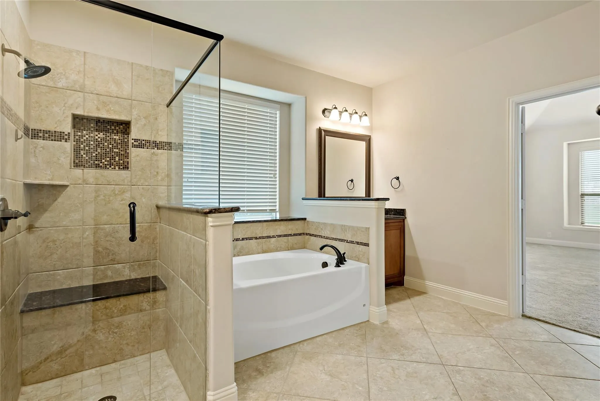 Spacious bathroom featuring a walk-in shower with a built-in bench and recessed shelf, a separate soaking tub, a vanity with a dark wood-finish cabinet and framed mirror, and large format tile flooring