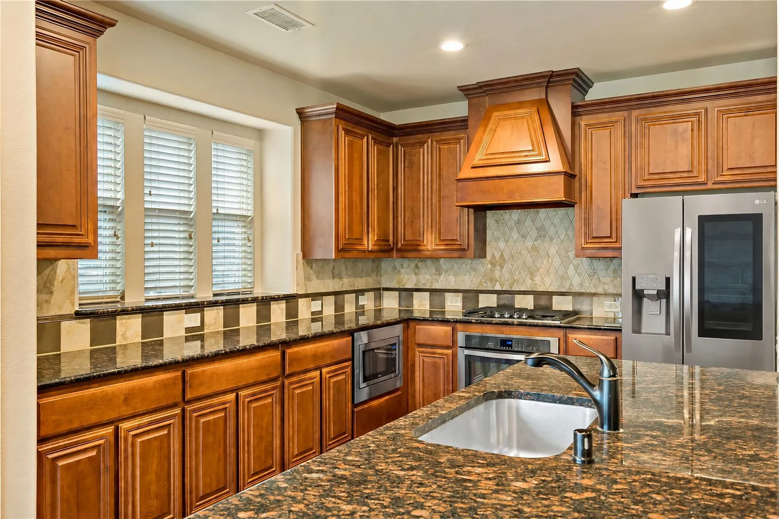 Kitchen featuring wood cabinetry, granite countertops, stainless steel appliances, a range hood, and a tile backsplash