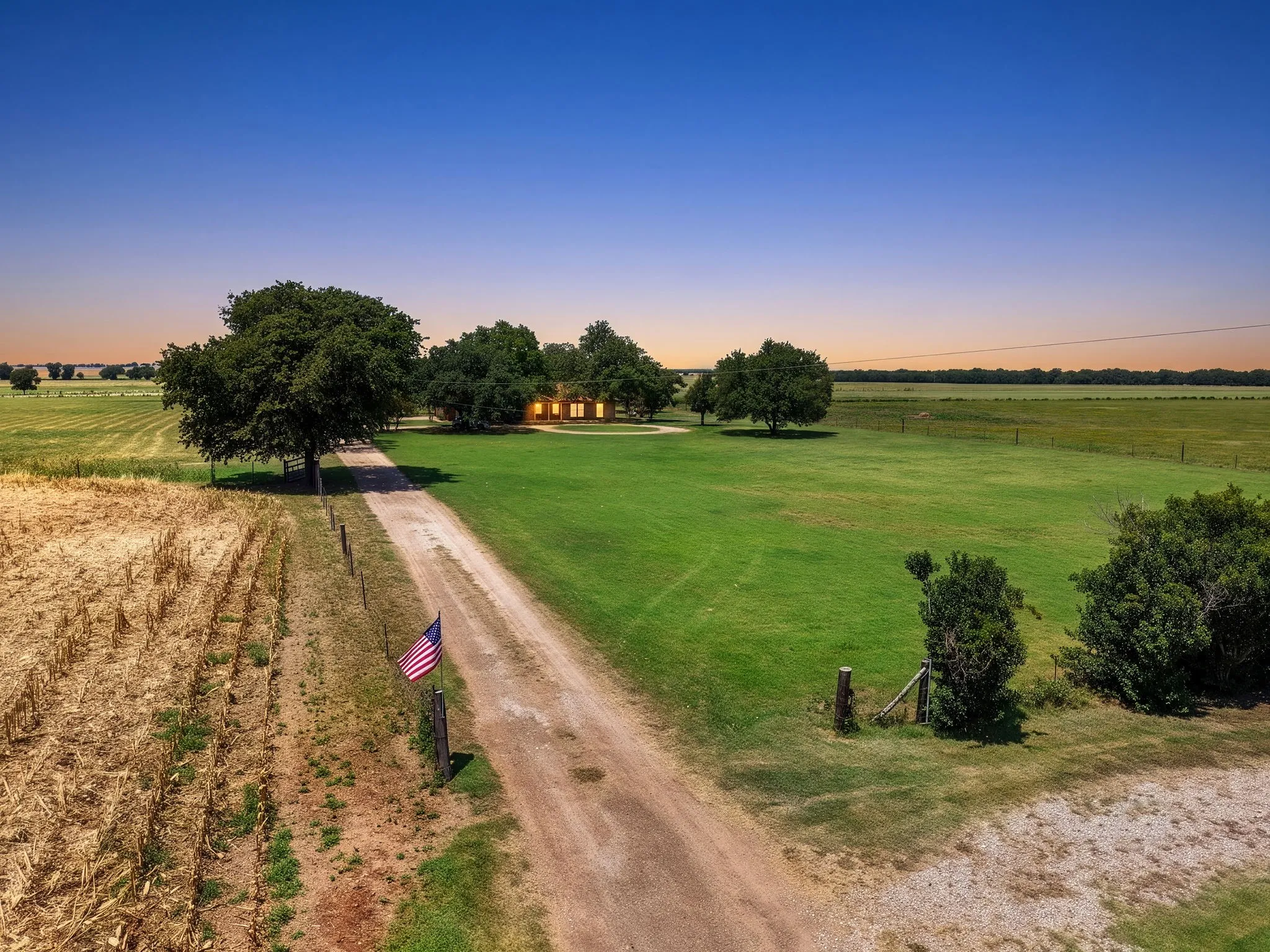 Expansive property featuring a long gravel driveway, a single-story residence surrounded by mature trees, and vast green fields
