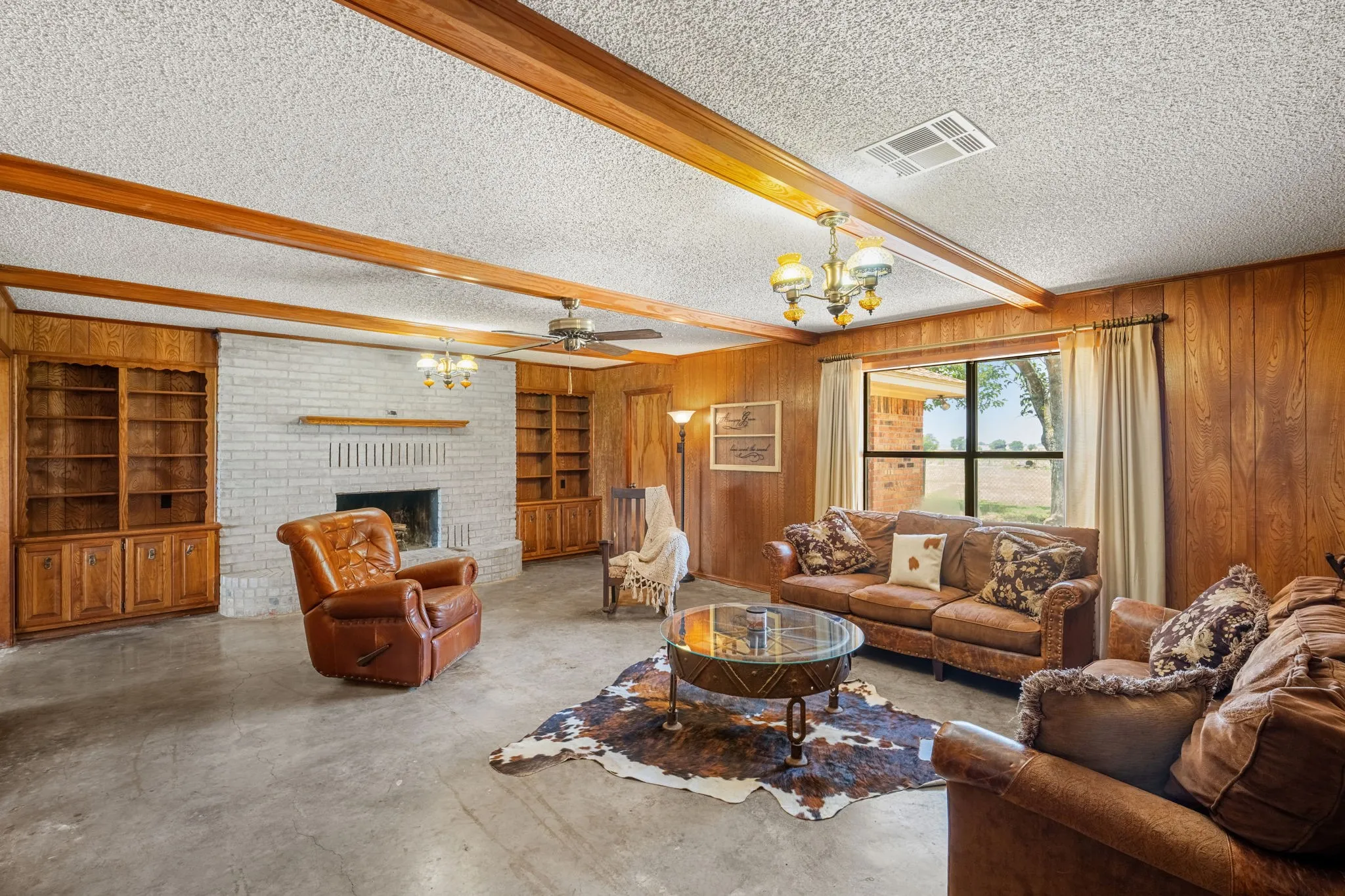 Spacious living area featuring exposed wood beams, light brick fireplace with wood mantle, concrete flooring, wood paneling, and built-in wood shelving units