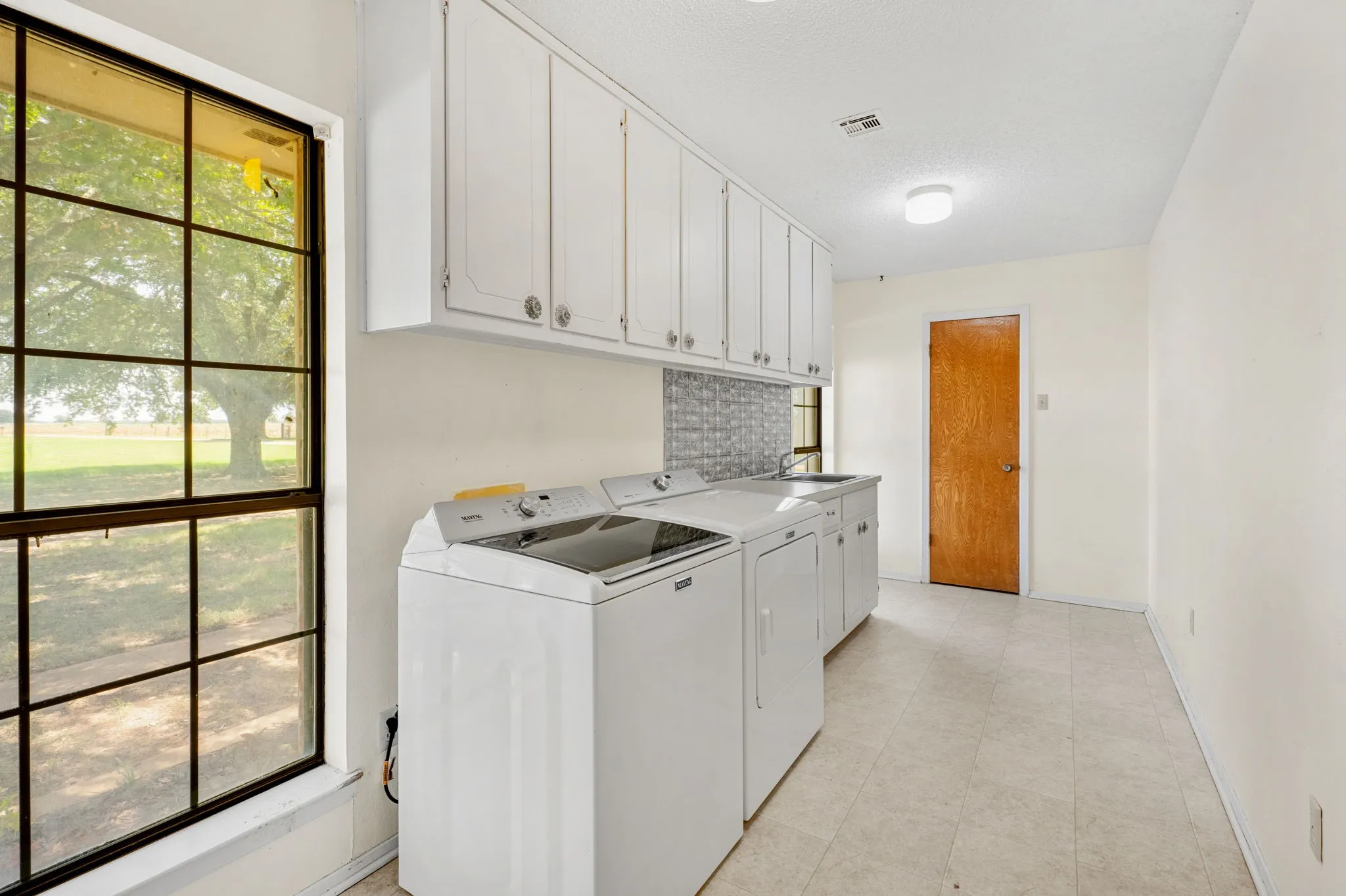 Utility area featuring a large grid-pane window, white upper cabinetry, a utility sink, and a grey tile backsplash