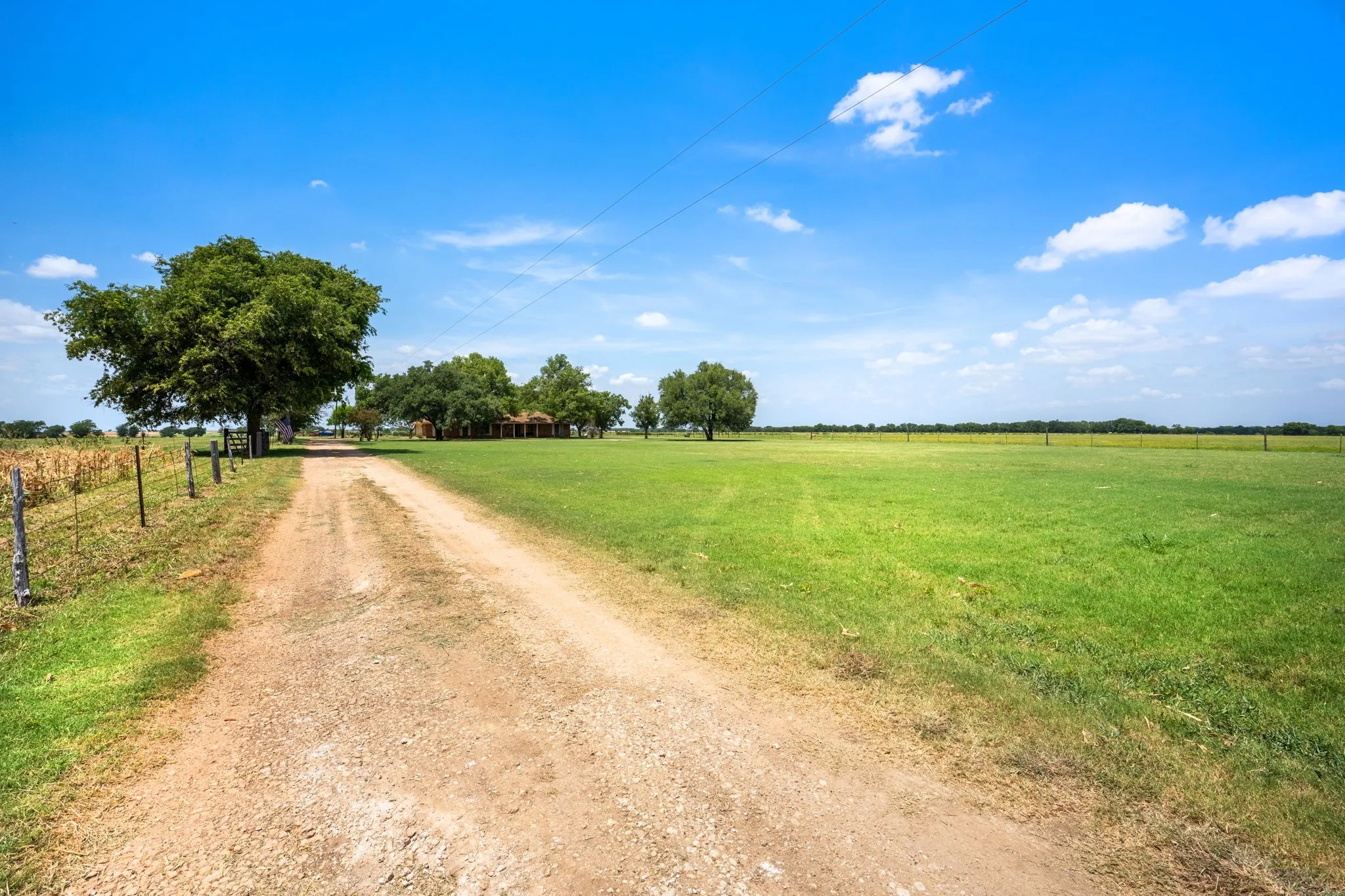 Gravel driveway leading to a residence, bordered by a wire fence and mature trees