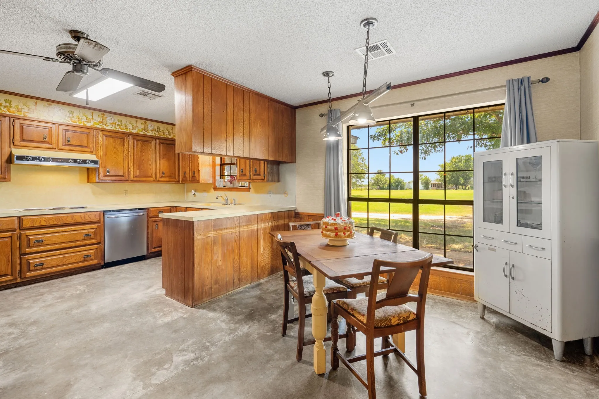Kitchen featuring wood cabinetry, stainless steel dishwasher, and a large window overlooking a landscaped area