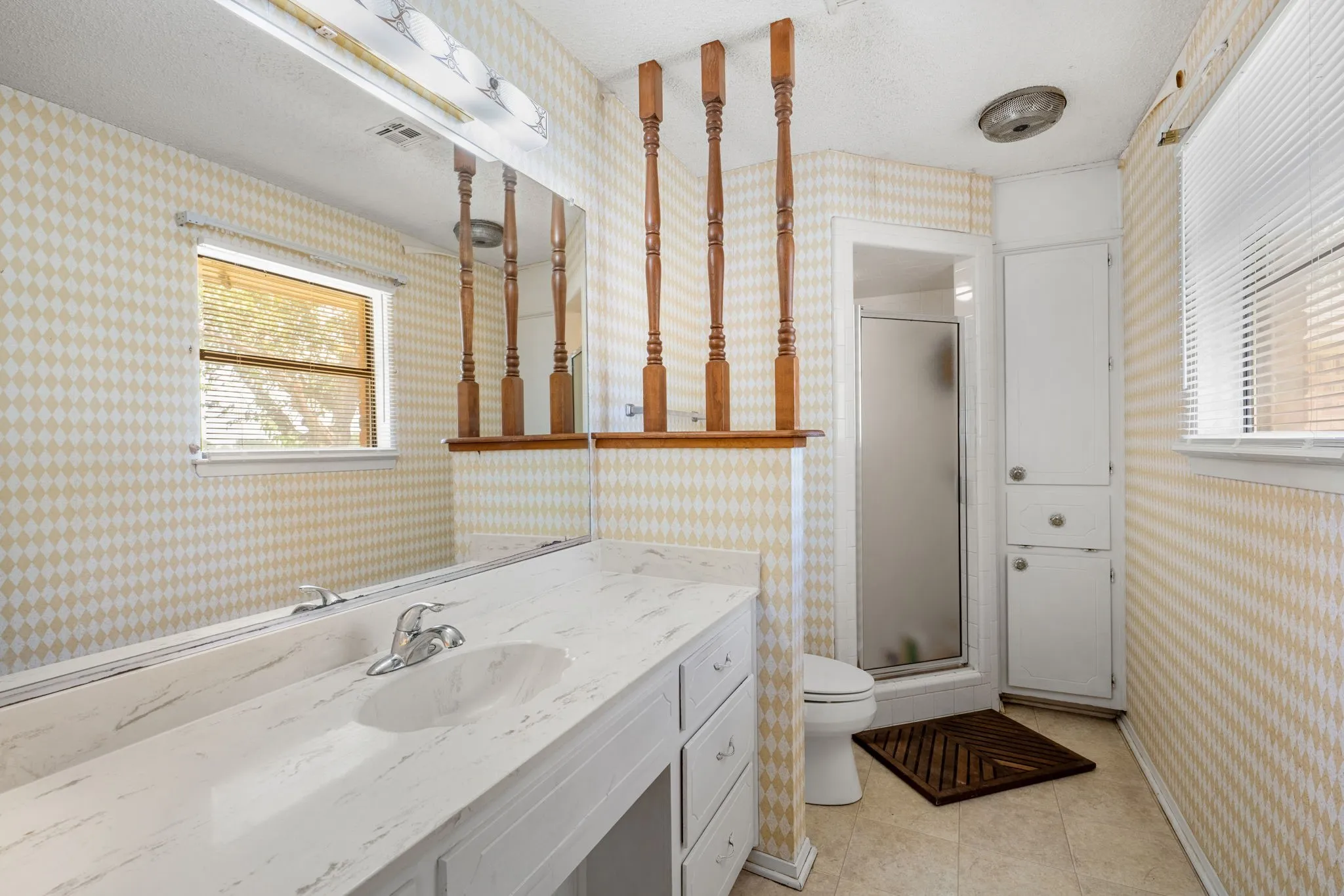 Bathroom featuring a single vanity with an integrated sink, patterned wallpaper, frosted glass shower enclosure, built-in storage cabinetry, and a window with blinds