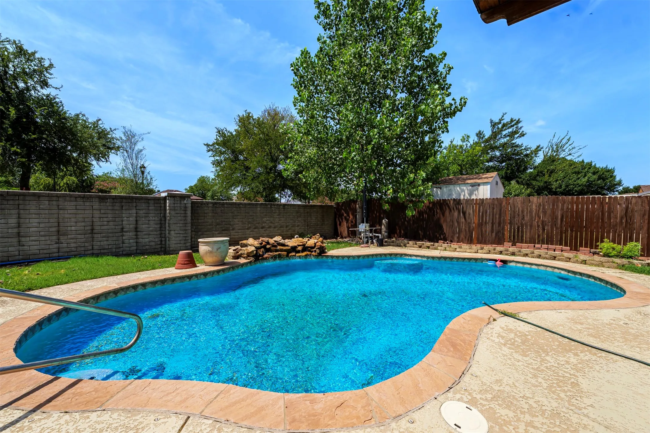 Freeform swimming pool with integrated rock waterfall feature, complemented by a perimeter of mature landscaping and a combination of block and wood fencing