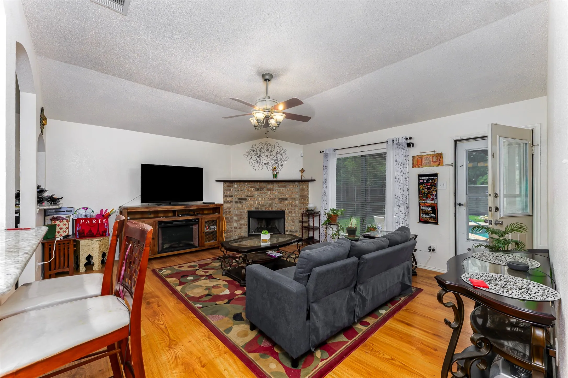 Living area featuring wood-finish flooring, a brick fireplace with a hearth, a ceiling fan, and a light-filled window overlooking outdoor space