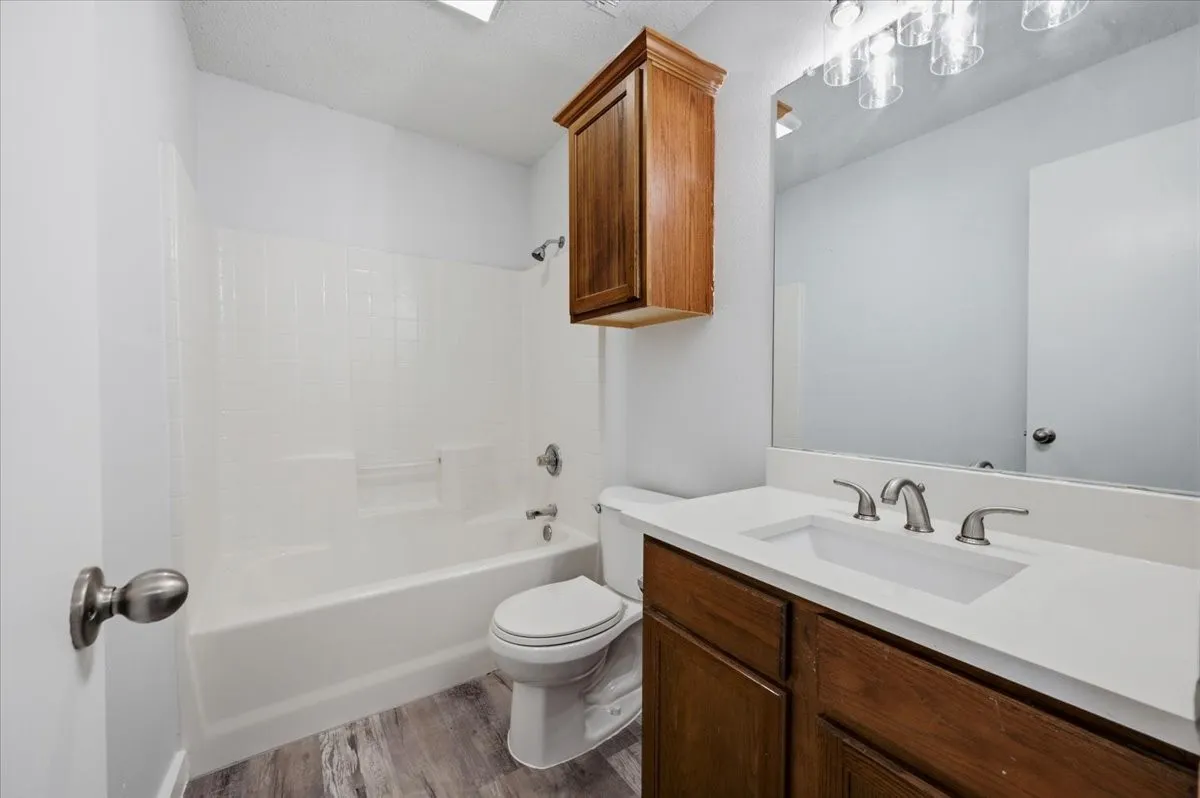 Bathroom featuring a white bathtub with an integrated shower, a white vanity countertop with an undermount sink, a wood-finish vanity cabinet, a wall-mounted mirror with overhead lighting, and wood-finish flooring