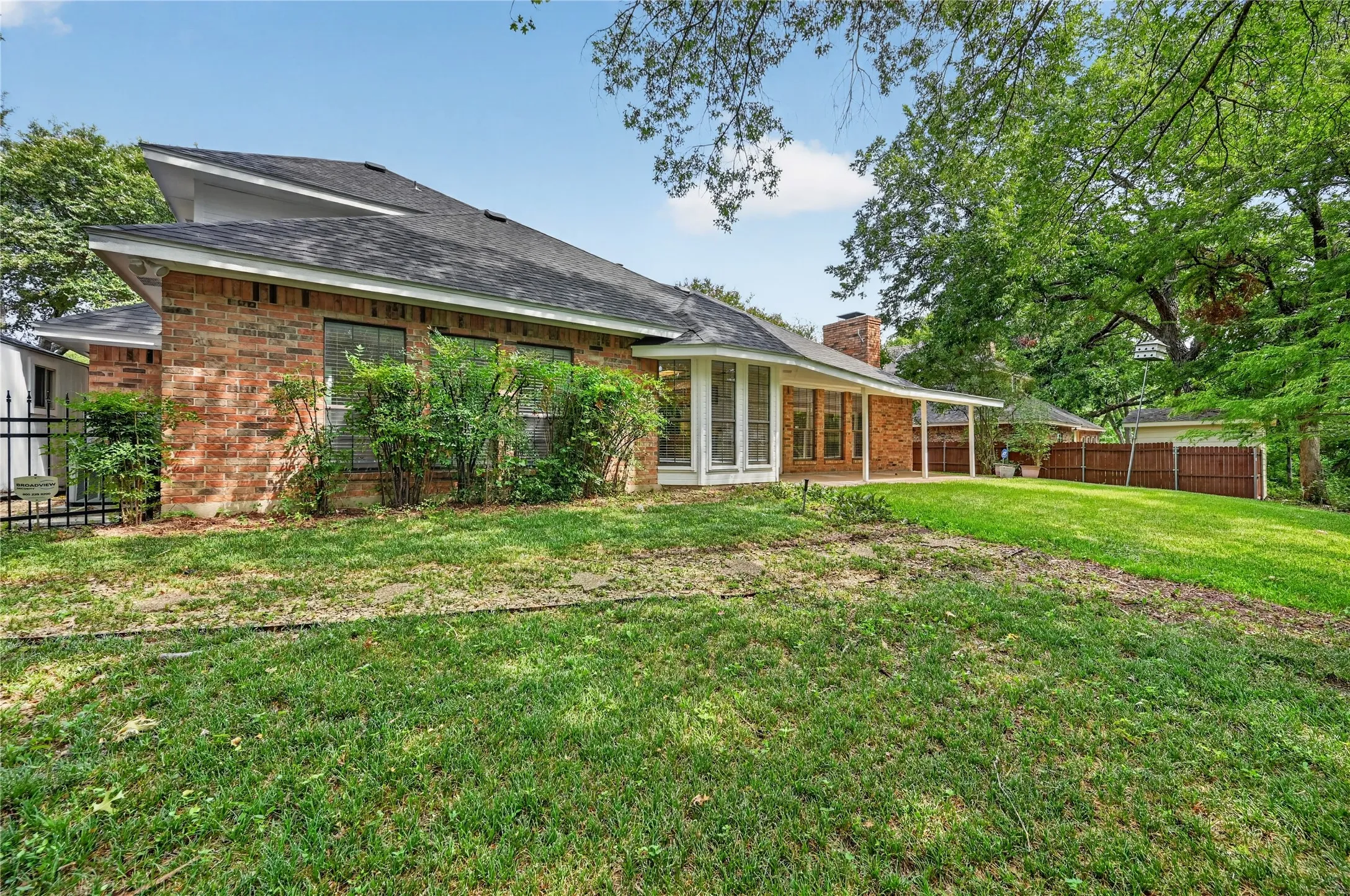 Expansive backyard featuring a brick exterior, covered patio, mature trees, and a privacy fence
