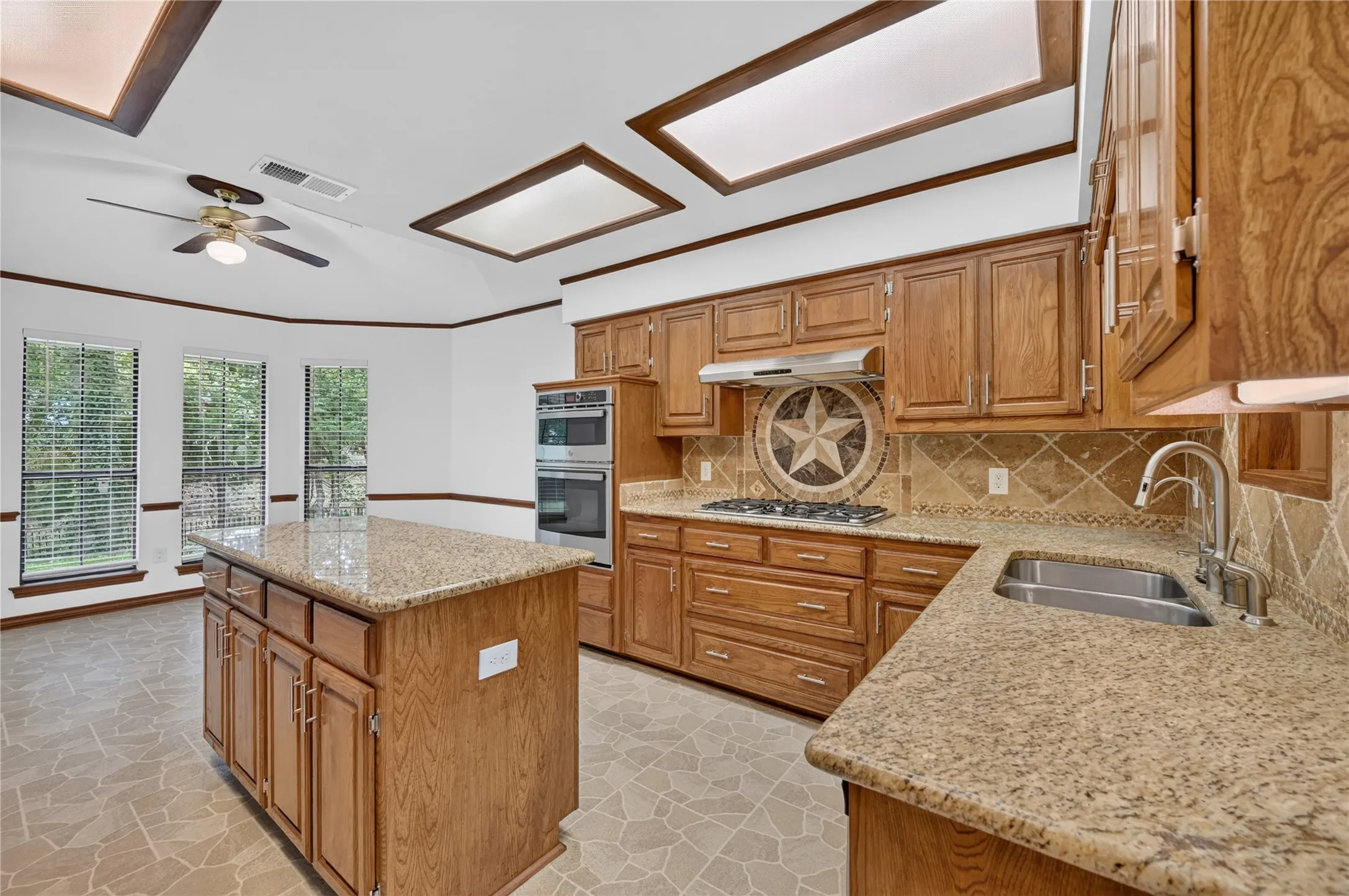 Kitchen featuring a central island with granite countertop, wood cabinetry, stainless steel double basin sink, and built-in double ovens