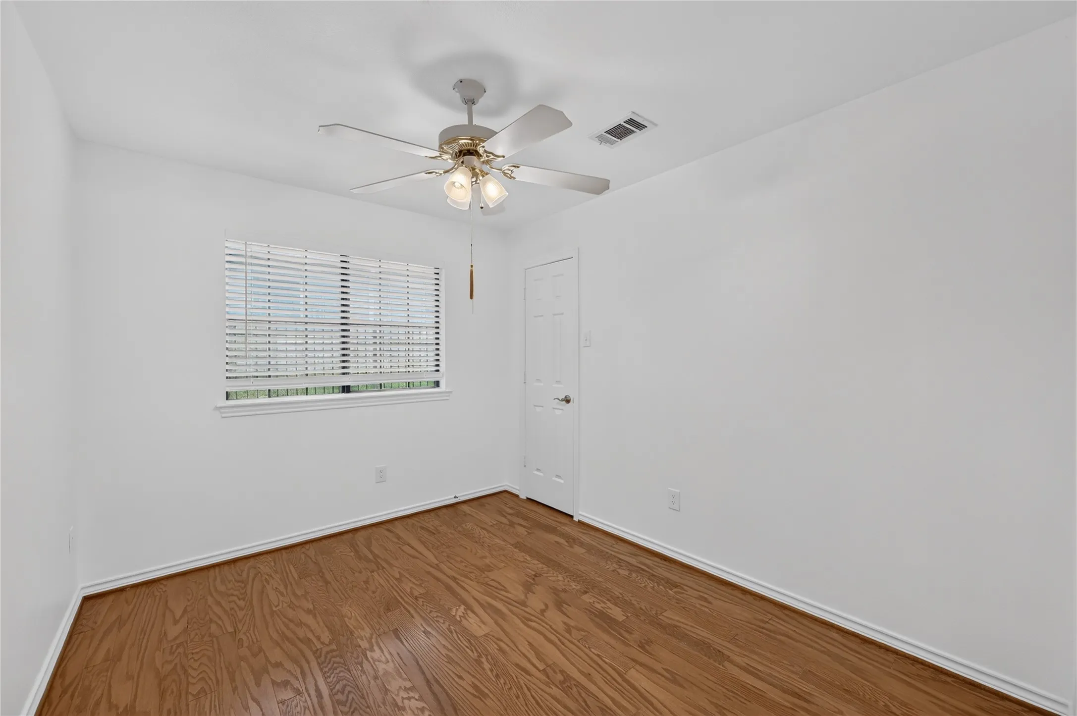Bright room featuring wood-finish flooring, a ceiling fan with integrated lighting, a single window with blinds, a white interior door, and white baseboards
