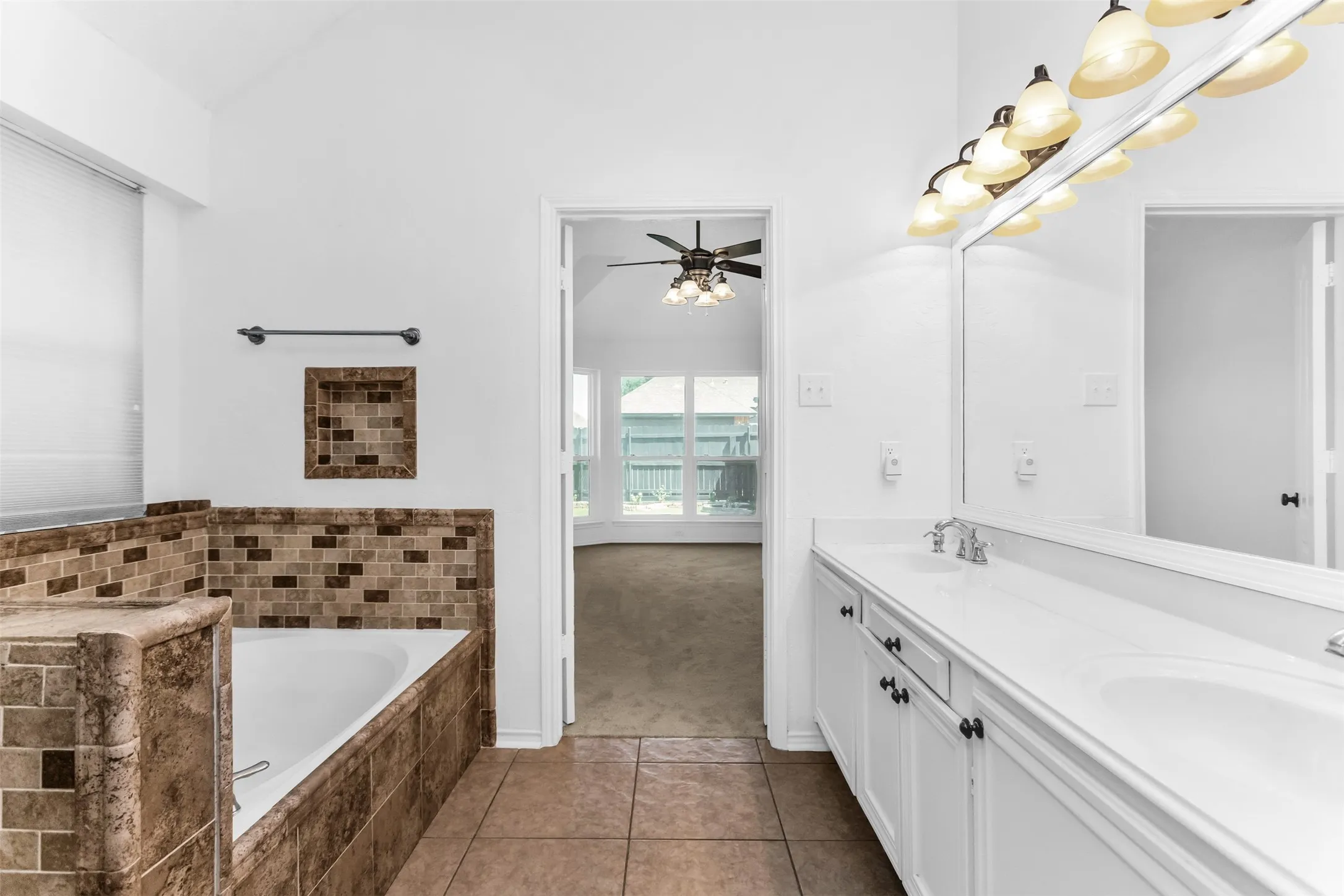 Tile flooring, built-in bathtub with tiled surround, dual vanity with white cabinets, and an expansive wall-mounted mirror with overhead lighting
