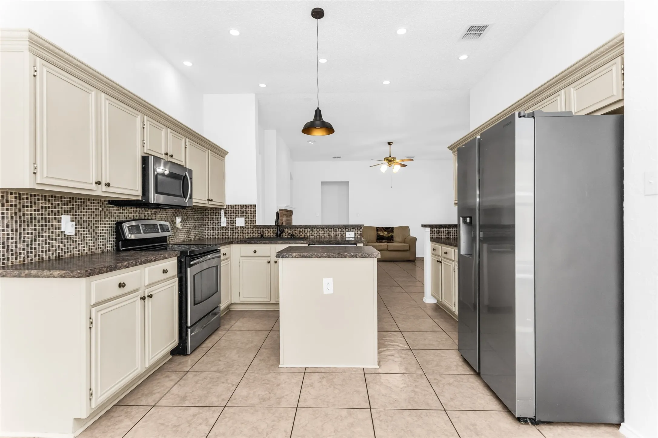Spacious kitchen featuring cream-colored cabinetry, stainless steel appliances, granite countertops, a large center island with seating, mosaic tile backsplash, tile flooring, and recessed ceiling lighting