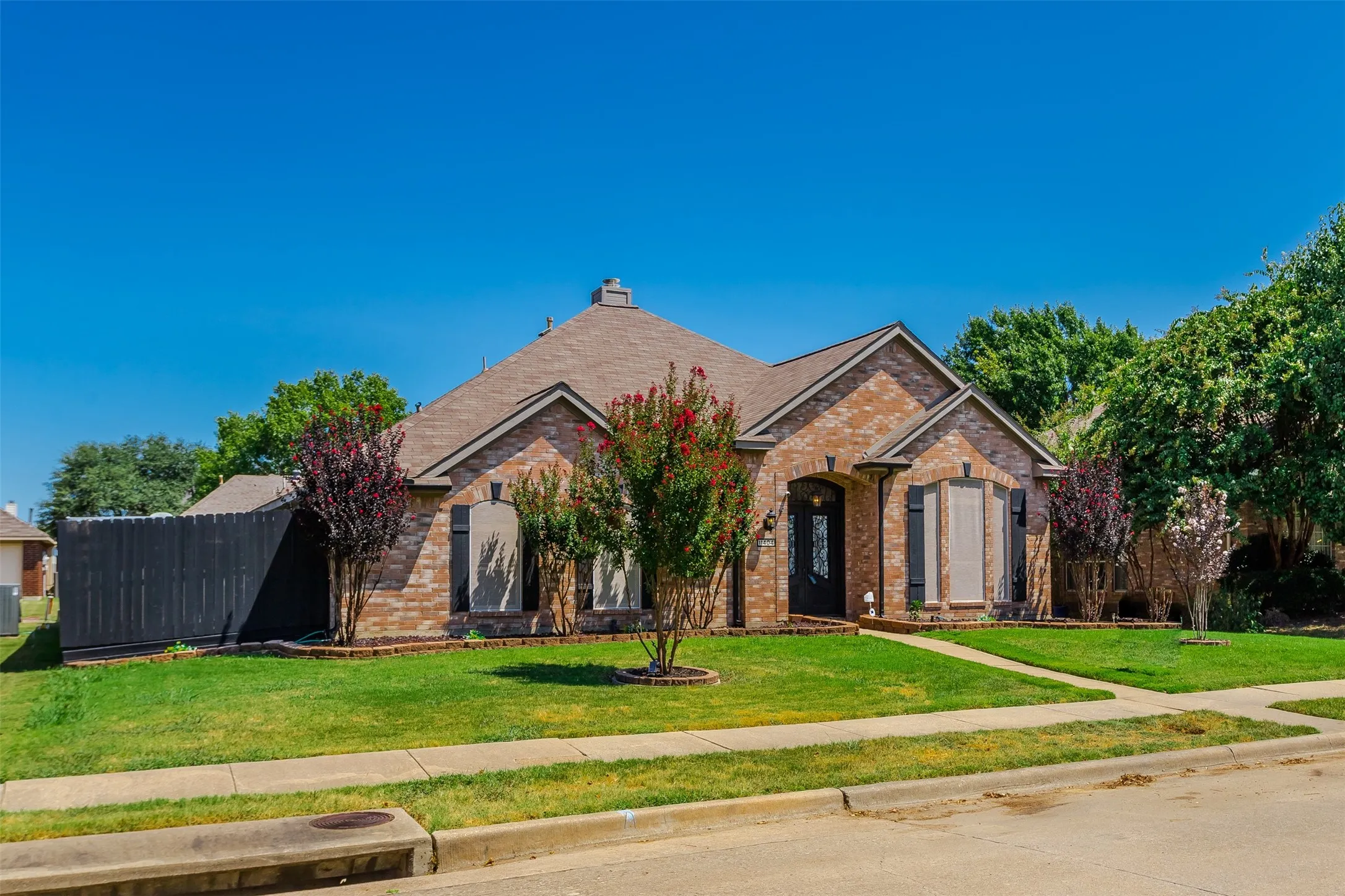Brick Colonial home showcasing a manicured front lawn, mature landscaping with flowering shrubs and trees, a gabled roofline with architectural detailing, shuttered windows, and a curved driveway approach