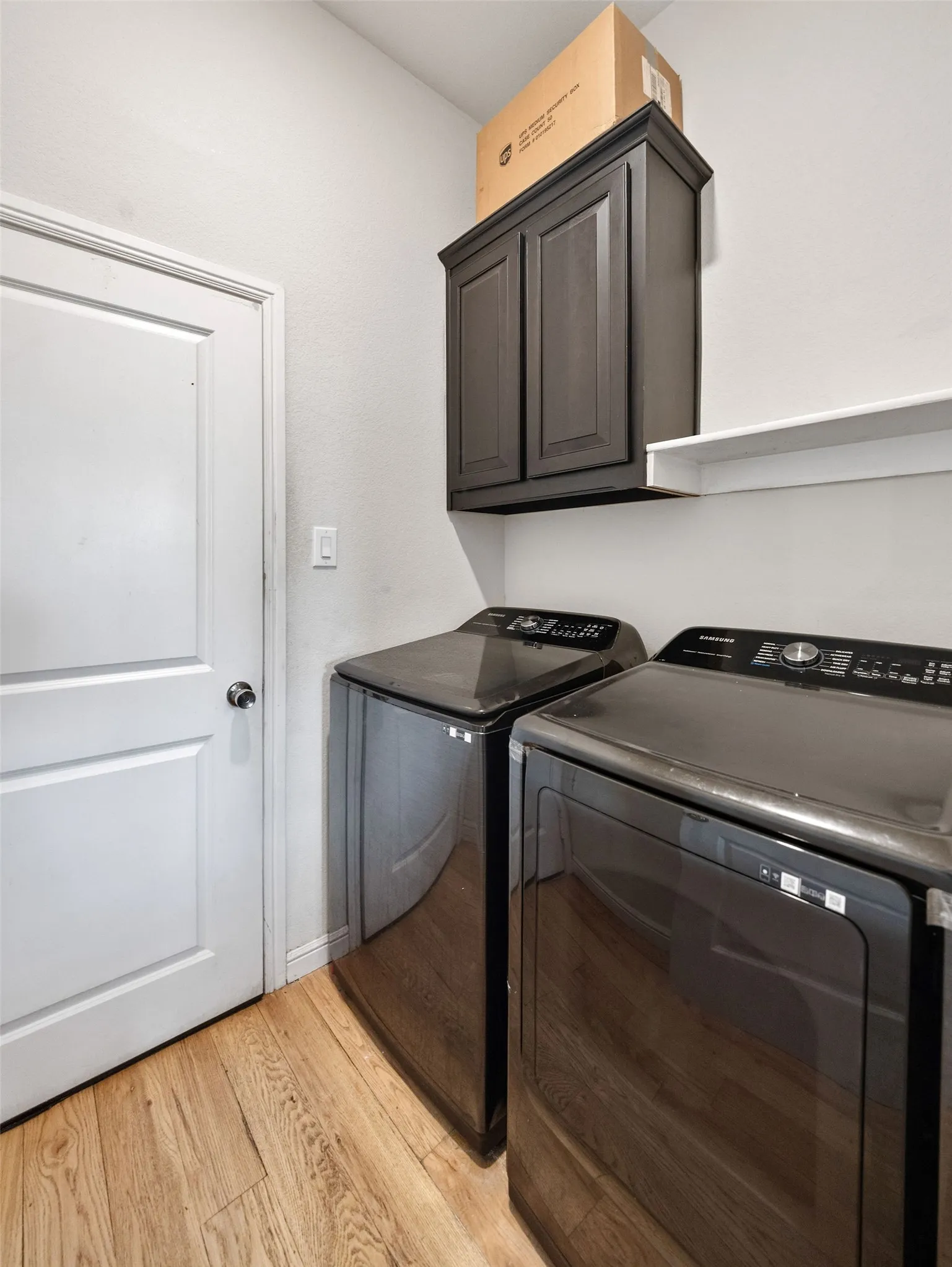 Laundry area featuring wood-finish flooring, a white interior door, a dark wood-finish wall cabinet, and a white wall shelf