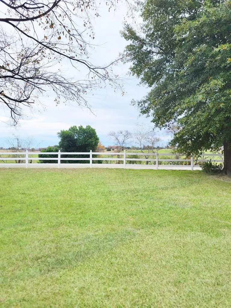 Spacious lawn area with a white picket fence, mature trees, and open-field backdrop
