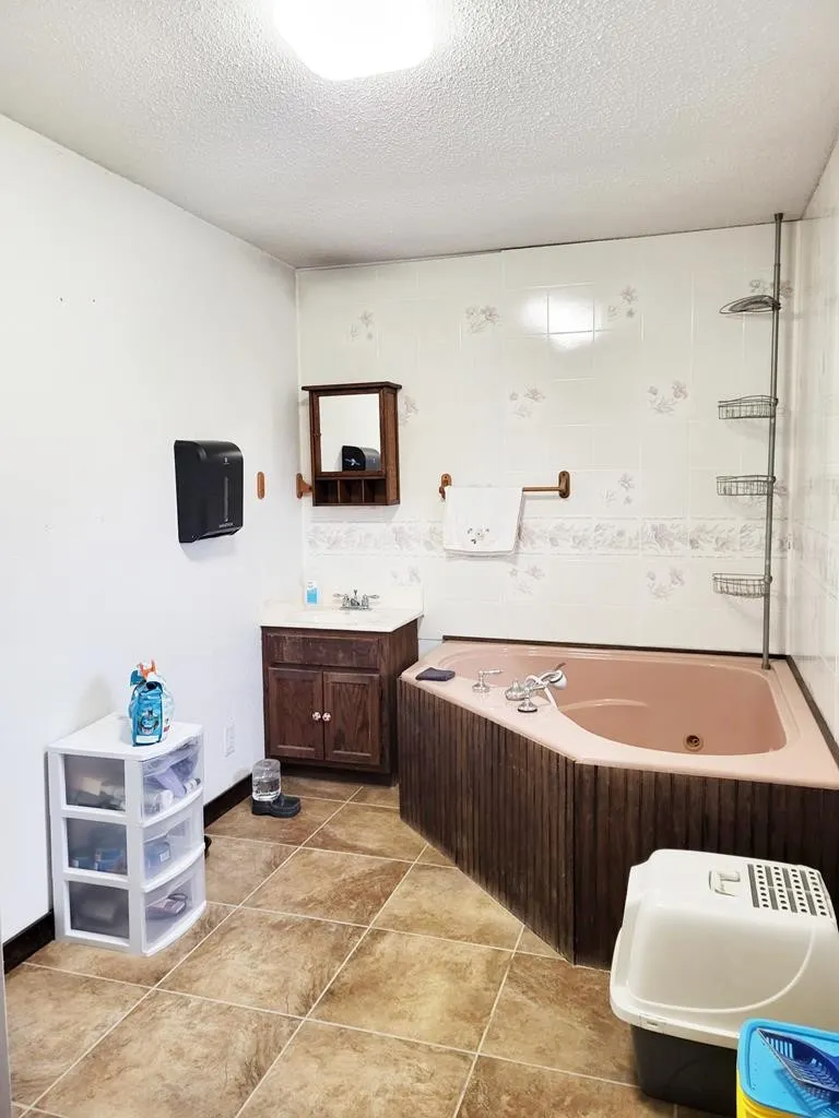Corner bathtub with wood-paneled surround, white tile walls with decorative accents, wall-mounted vanity mirror with shelf, and tile flooring
