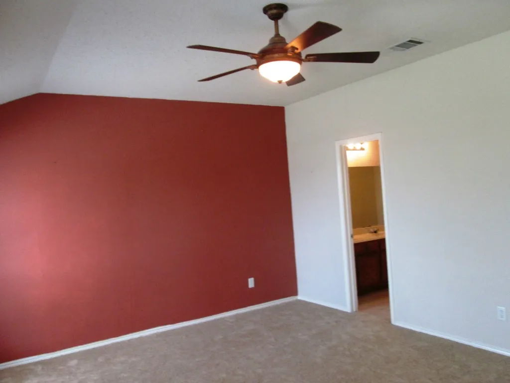 Spacious room with a ceiling fan light fixture, red accent wall, neutral carpeting, and white baseboards