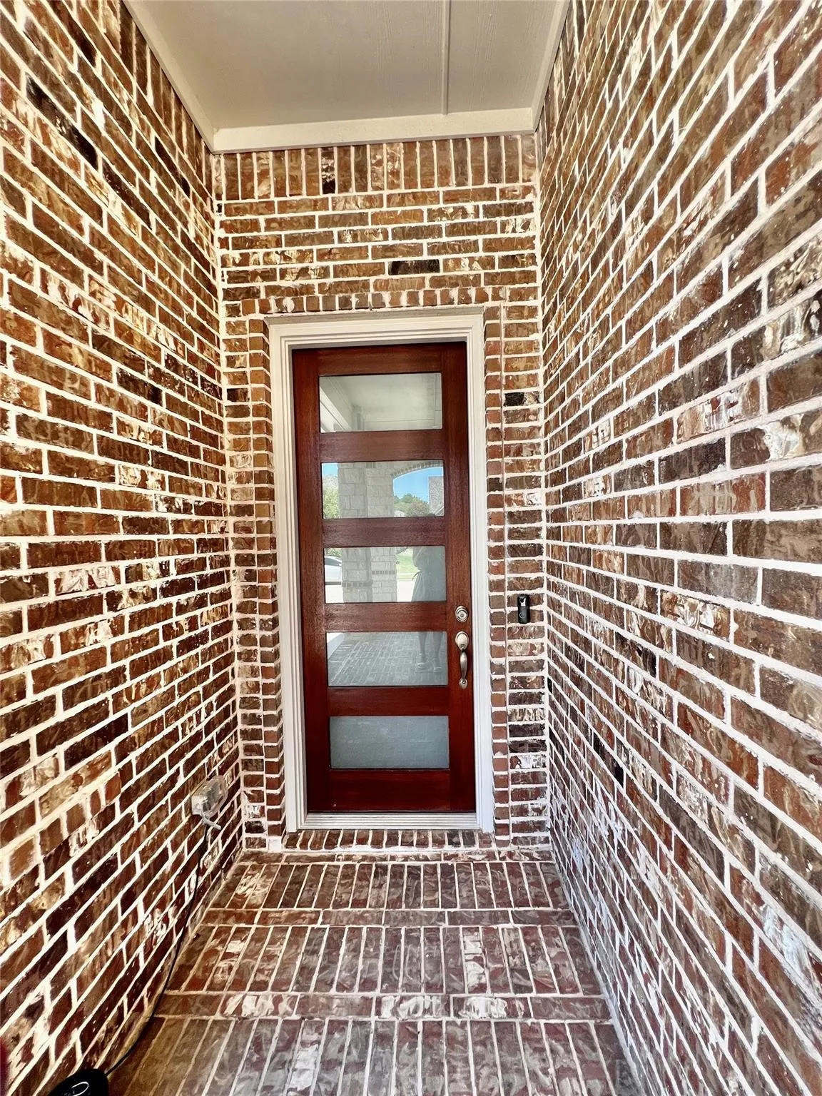 Brick-clad entryway featuring a rich wood-finish door with frosted glass panels, white trim, and a brick-patterned porch floor