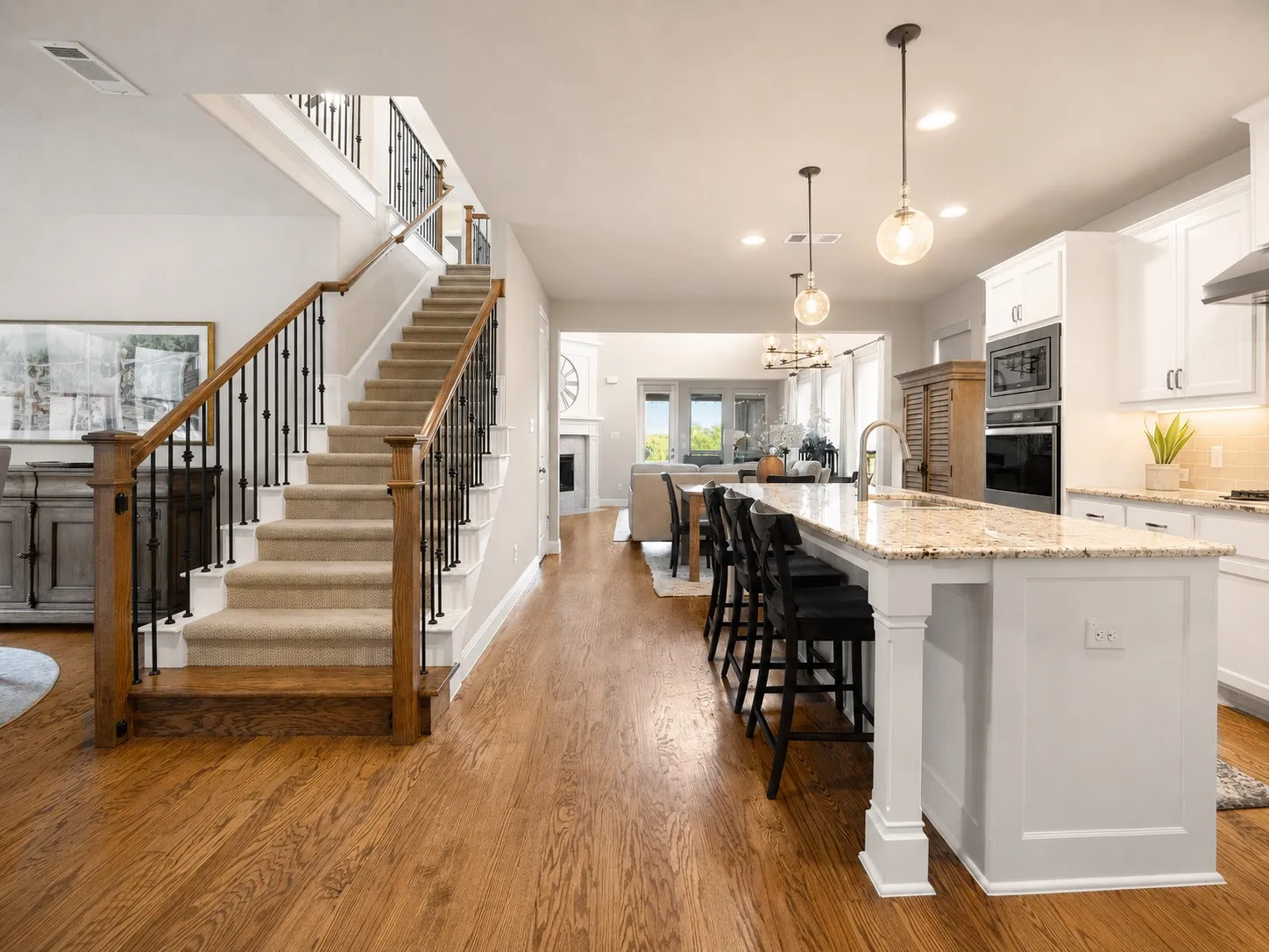 Open-concept interior featuring wood-finish flooring, a carpeted staircase with wood and wrought iron railings, a kitchen island with stone countertop, white cabinetry, and built-in stainless steel appliances