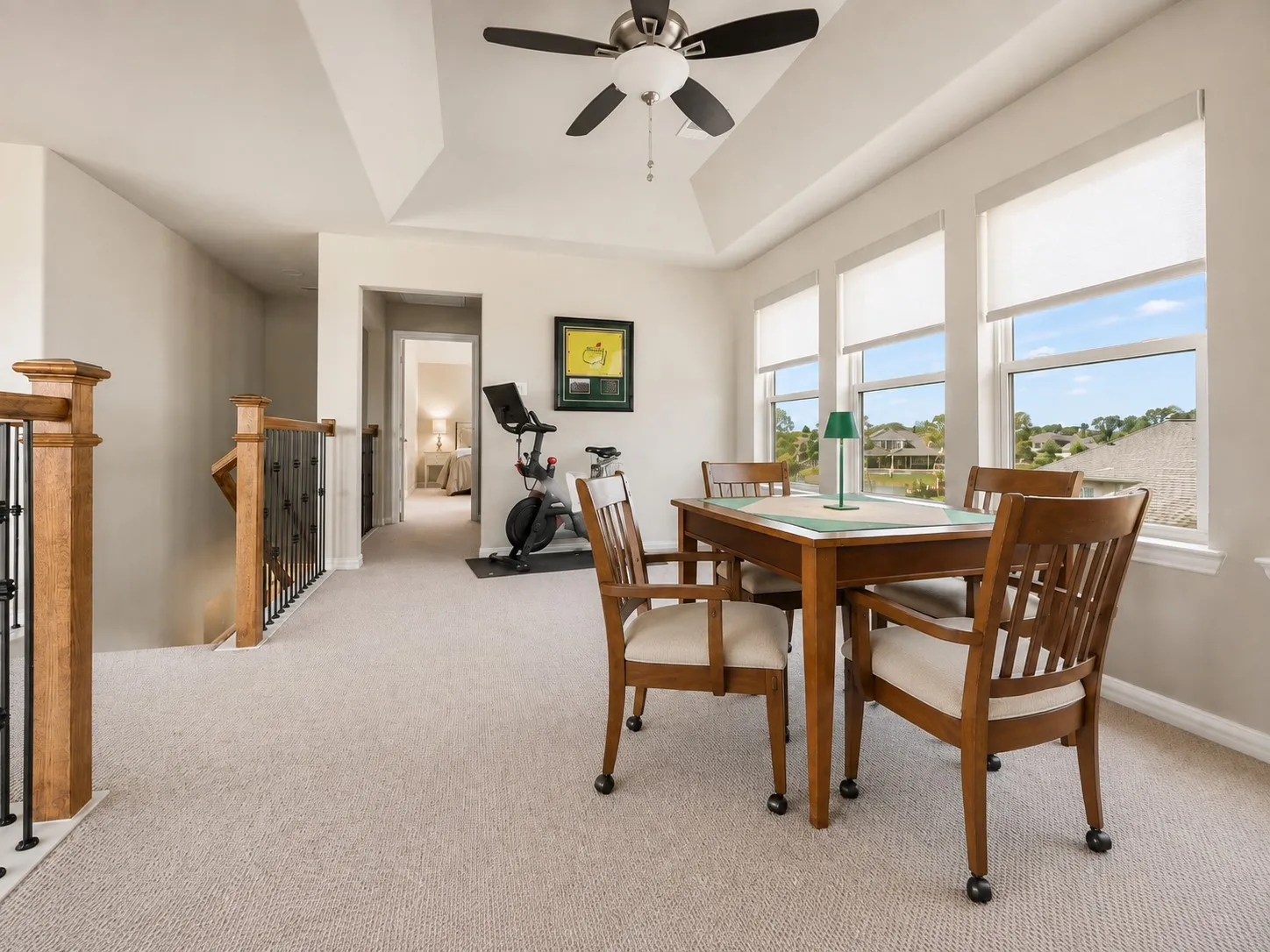 Carpeted loft area featuring a tray ceiling, ceiling fan, and multiple windows with roller shades