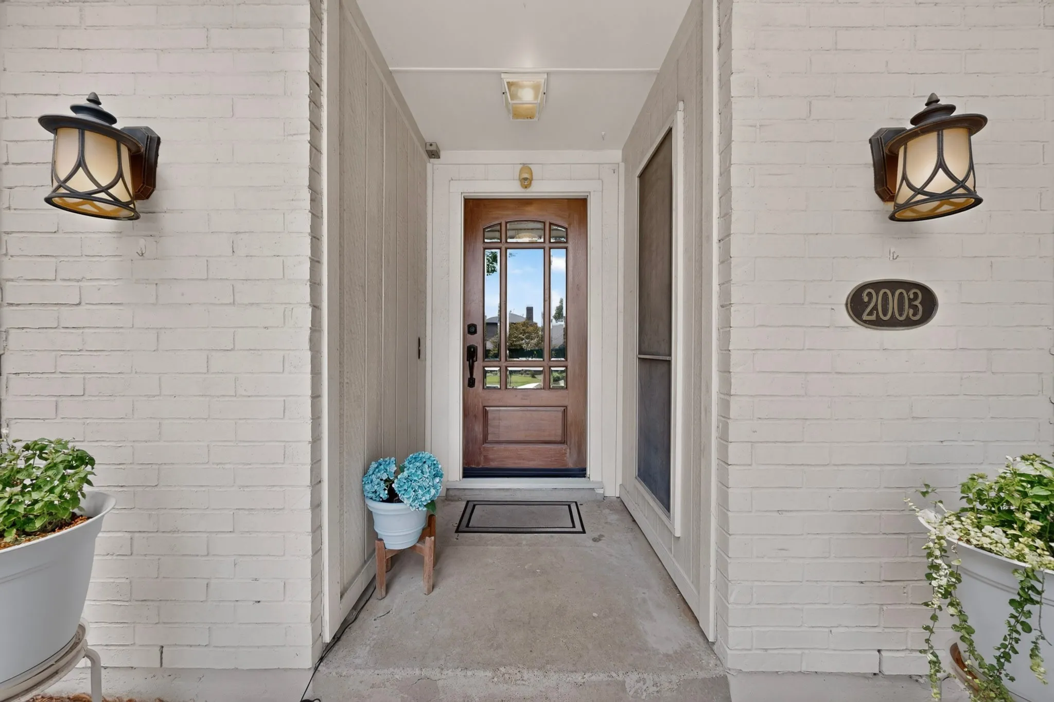 Welcoming entry featuring a wood-finish door with decorative glass, two wall-mounted lanterns, a concrete entry porch, and a white brick exterior