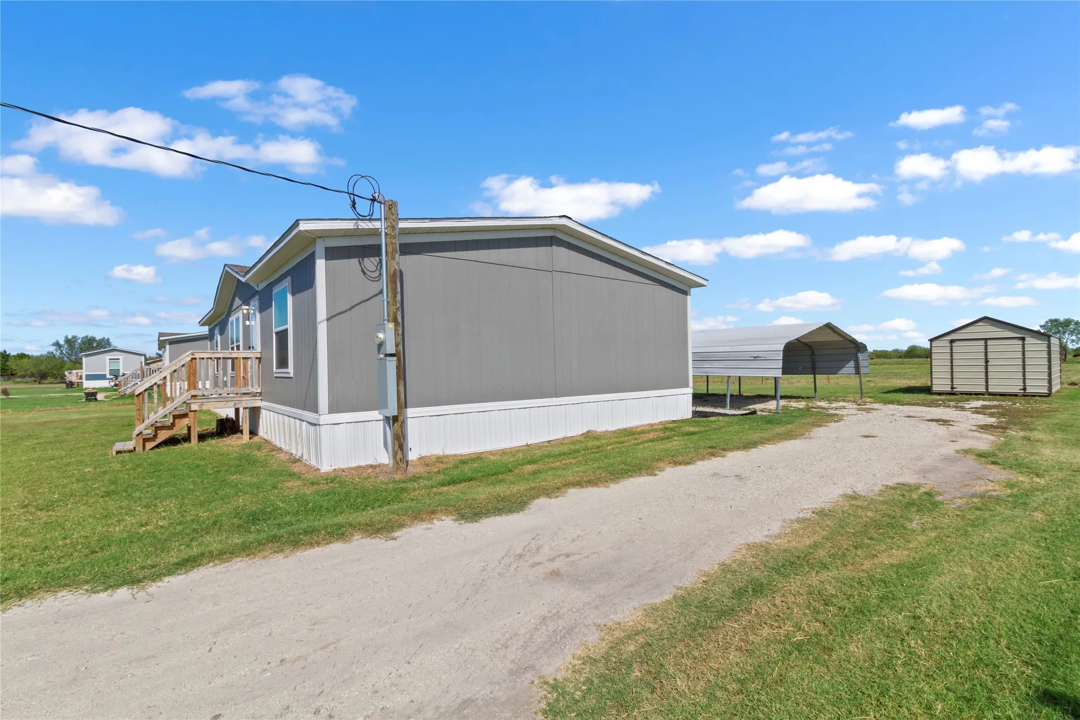 Gray-sided manufactured home featuring white trim, a wooden porch with steps, a gravel driveway, a metal carport, and a separate storage shed