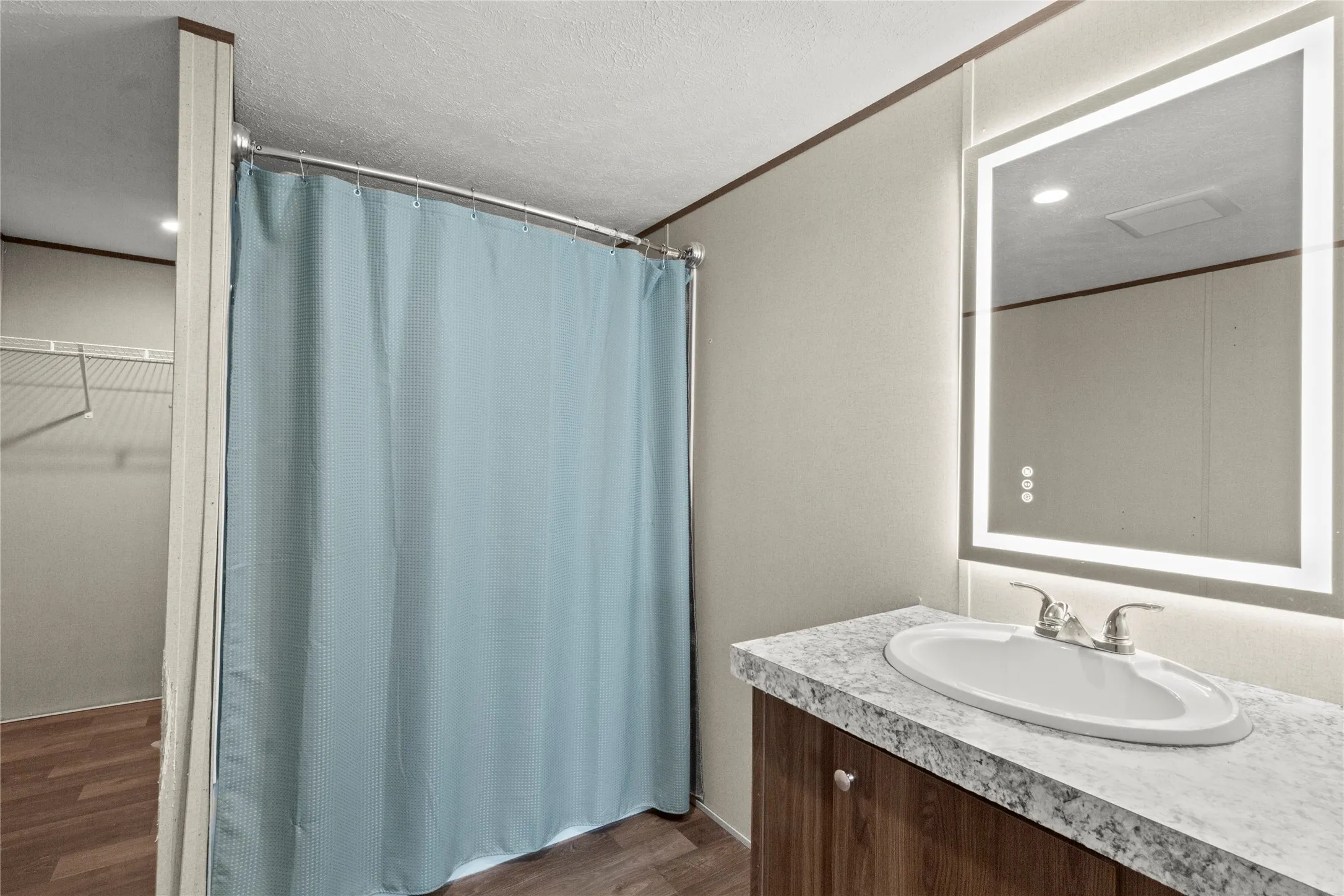 Bathroom featuring a vanity with an integrated sink, a lighted mirror, wood-finish flooring, a shower curtain, and a wall-mounted wire shelf