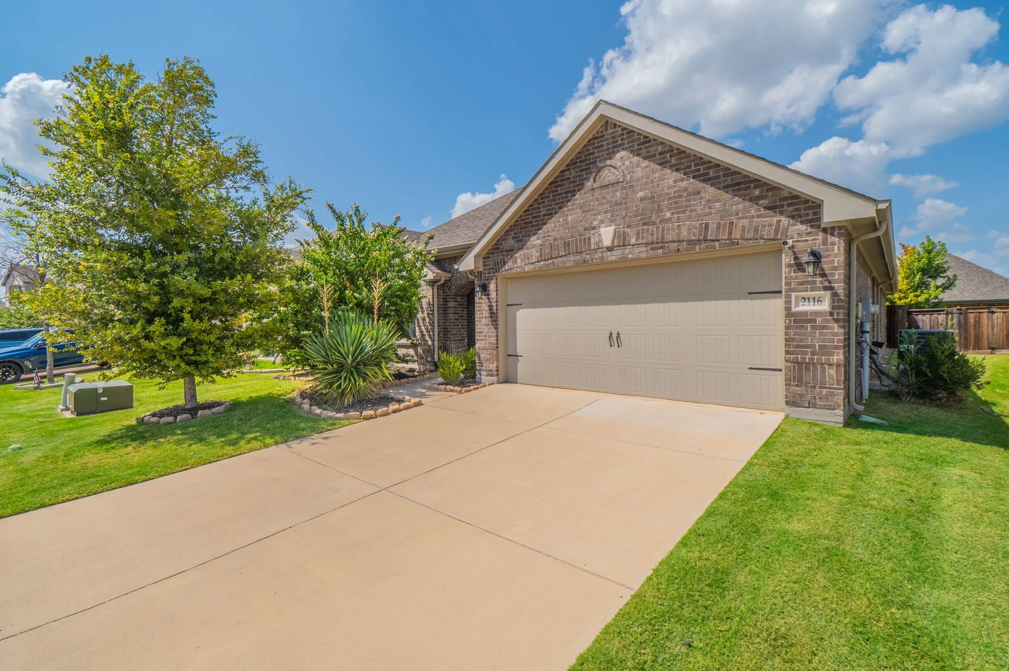 Brick exterior featuring a two-car garage with a panel door, concrete driveway, and front landscaping with mature trees and shrubs