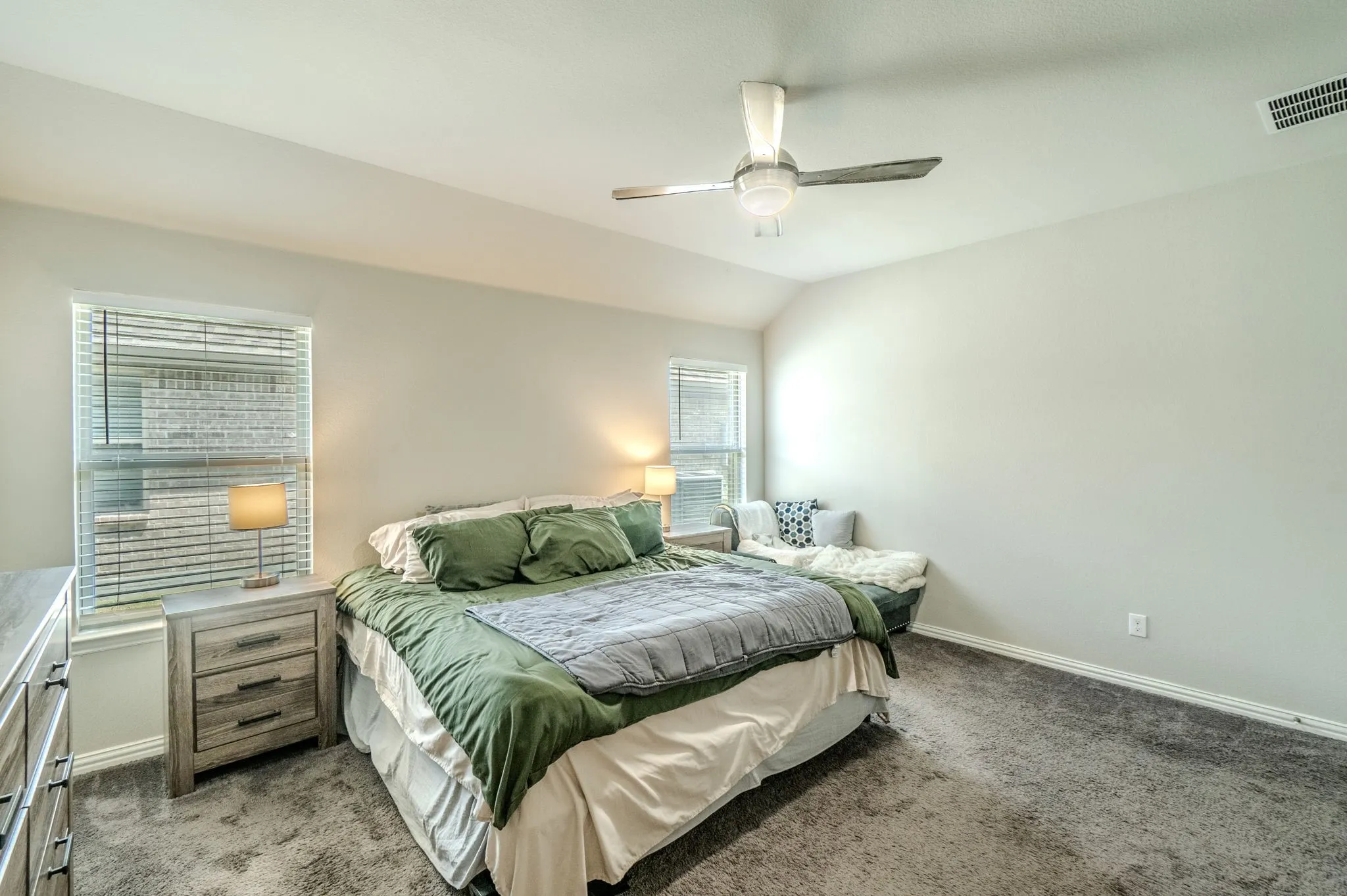 Carpeted room featuring a ceiling fan, vaulted ceiling, two windows with blinds, and light-colored walls