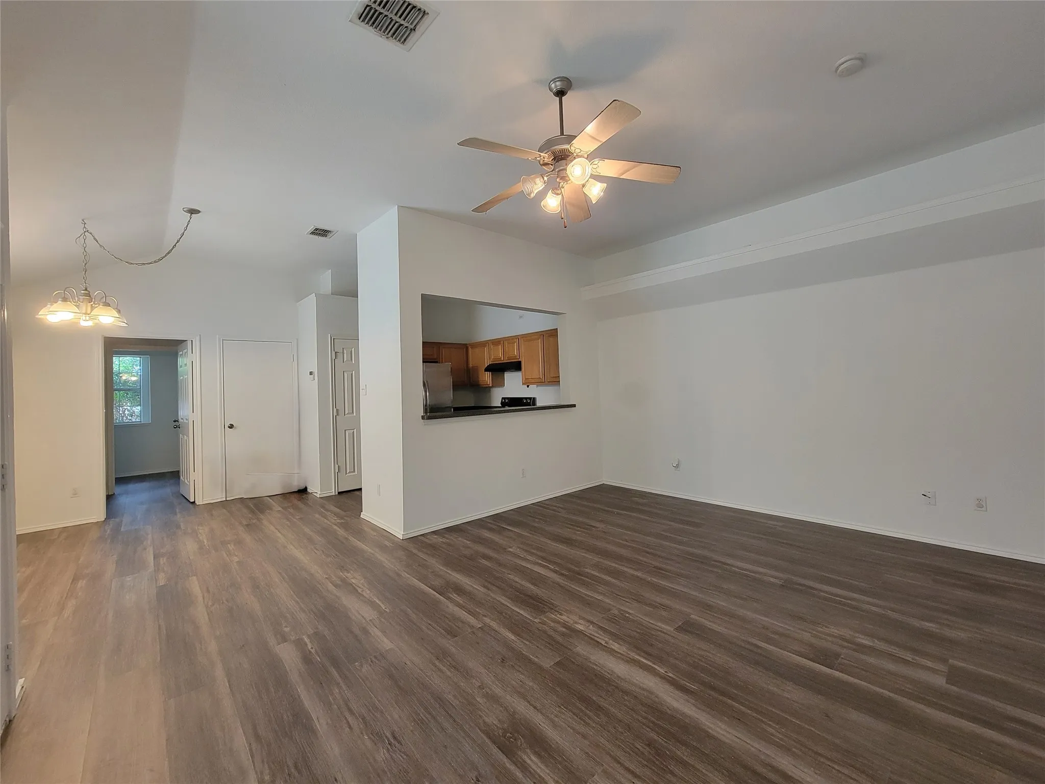 Open concept living area featuring wood-finish flooring throughout, a ceiling fan with integrated lighting, and a pass-through kitchen with wood cabinetry and a dark countertop