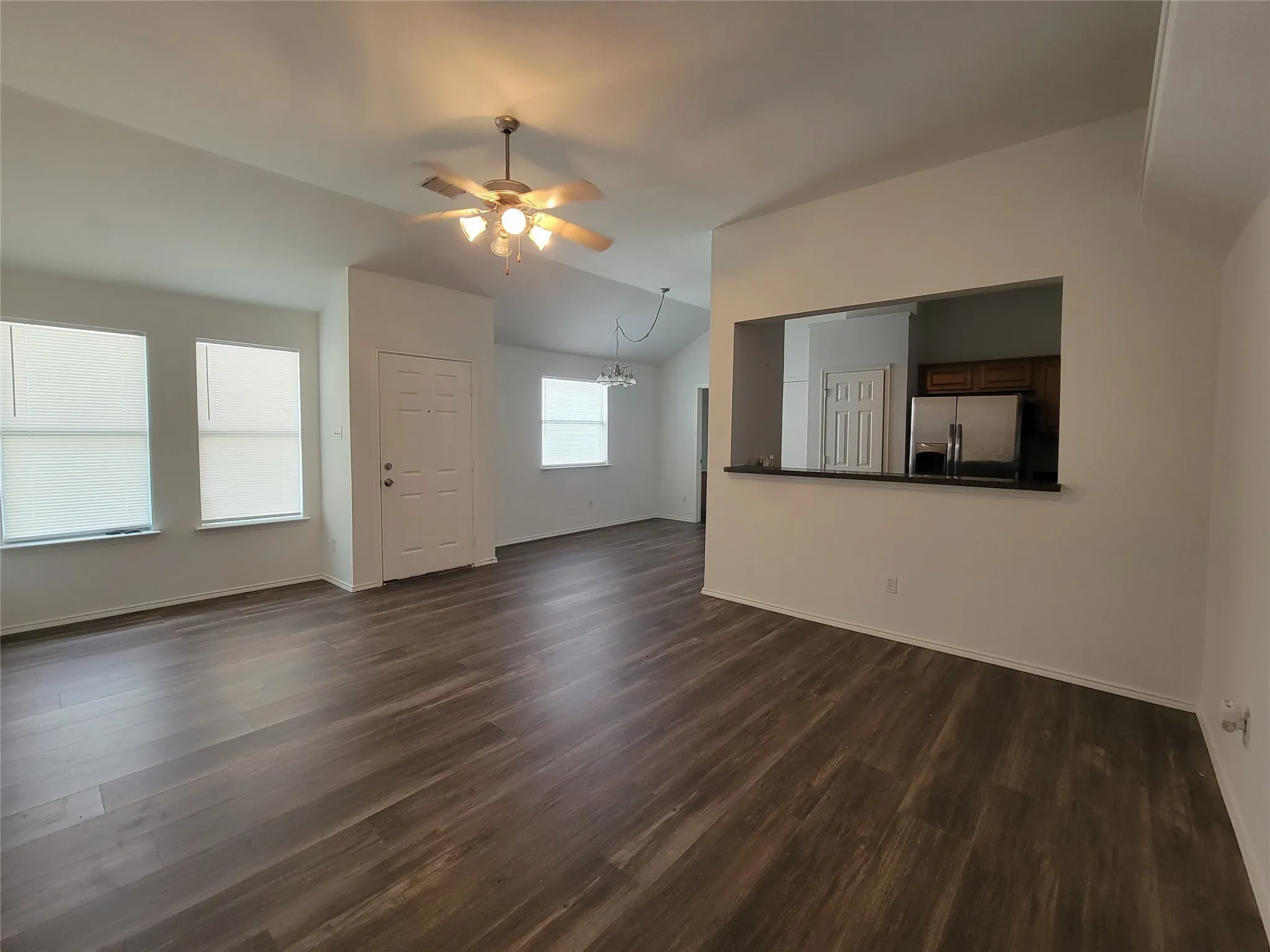 Spacious living area featuring wood-finish flooring, a ceiling fan with integrated lighting, multiple windows with blinds, a white paneled door, and a kitchen pass-through with a dark countertop