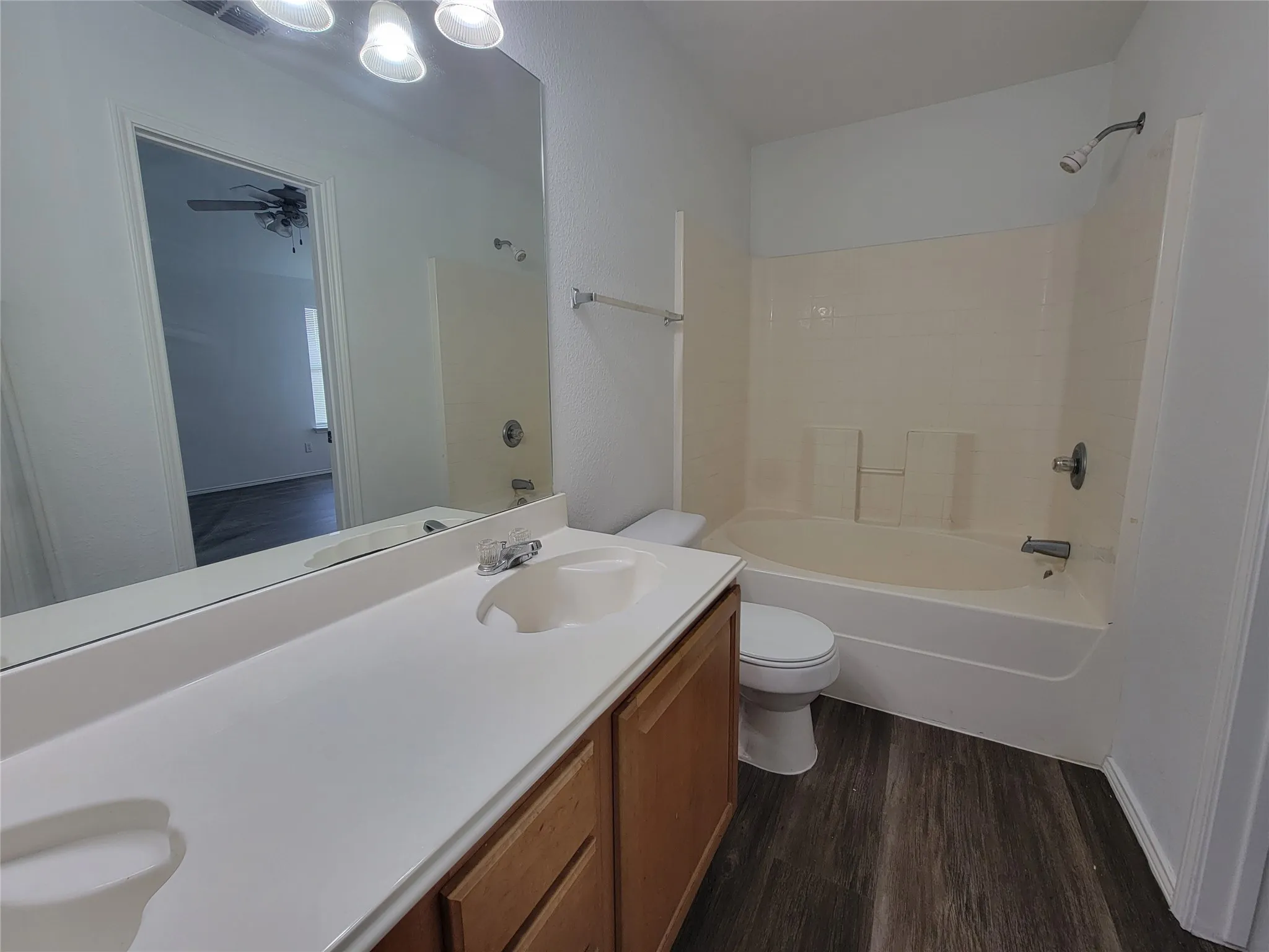 Bathroom featuring a wood-finish vanity with a white countertop and integrated sink, a rectangular wall-mounted mirror, a tub/shower combination with light-colored tile surround, and wood-finish flooring