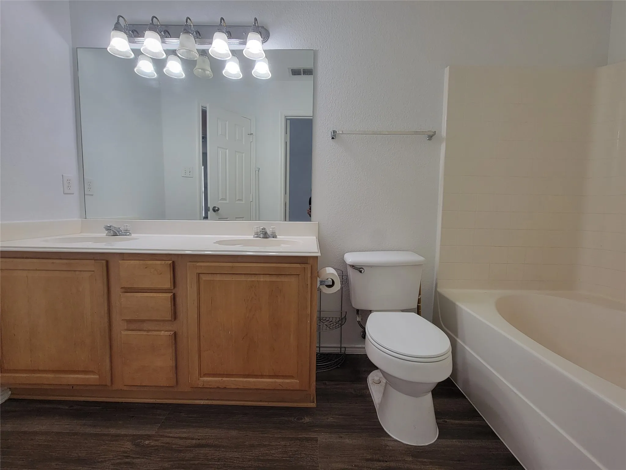 Bathroom featuring a double vanity with light wood cabinetry, a large mirror, a multi-bulb vanity light fixture, a toilet, and a bathtub with an integrated shower surround