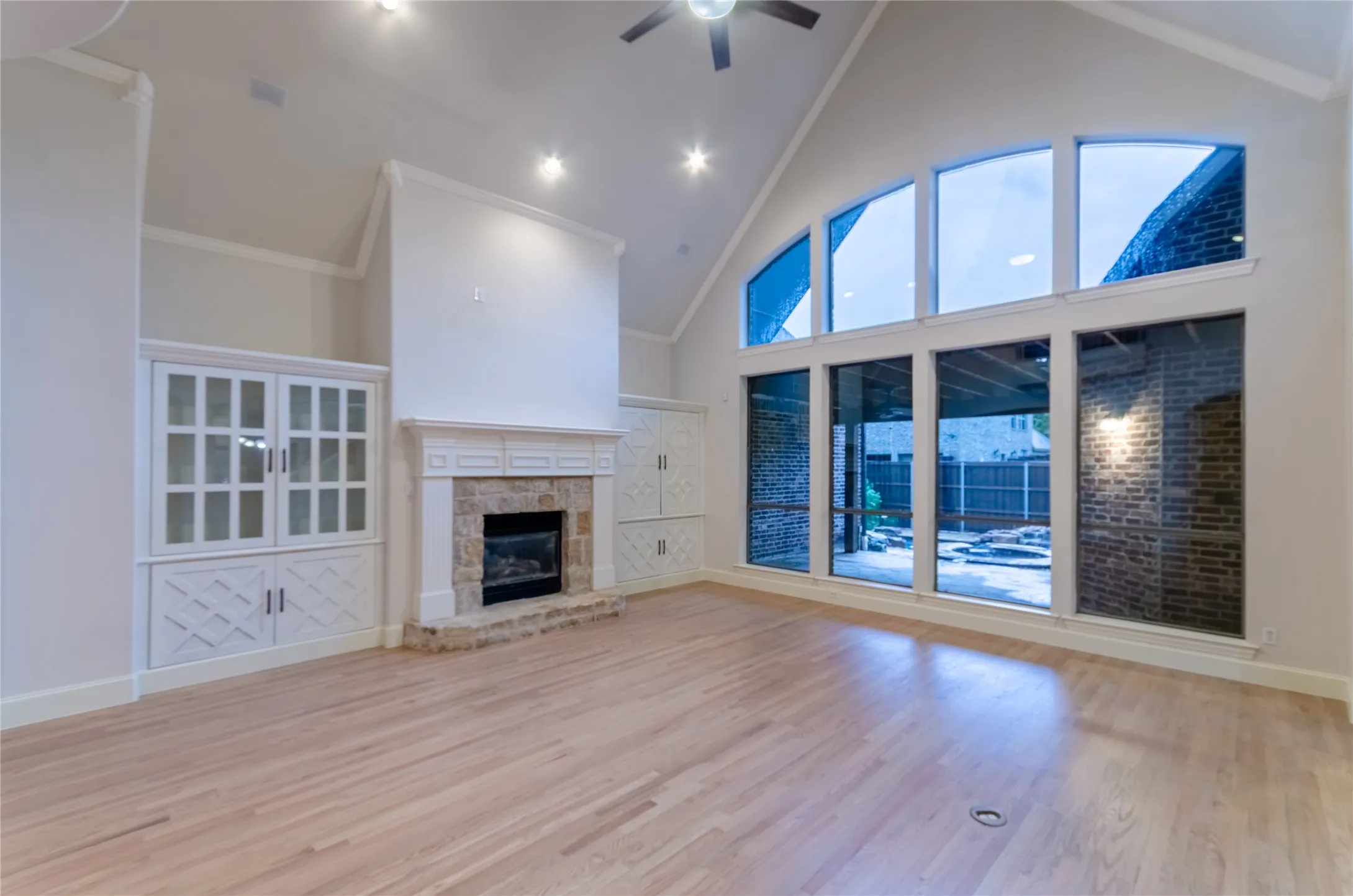 Vaulted ceiling great room featuring oversized windows, light wood-finish flooring, a stone-surround fireplace with a mantel, and built-in cabinetry