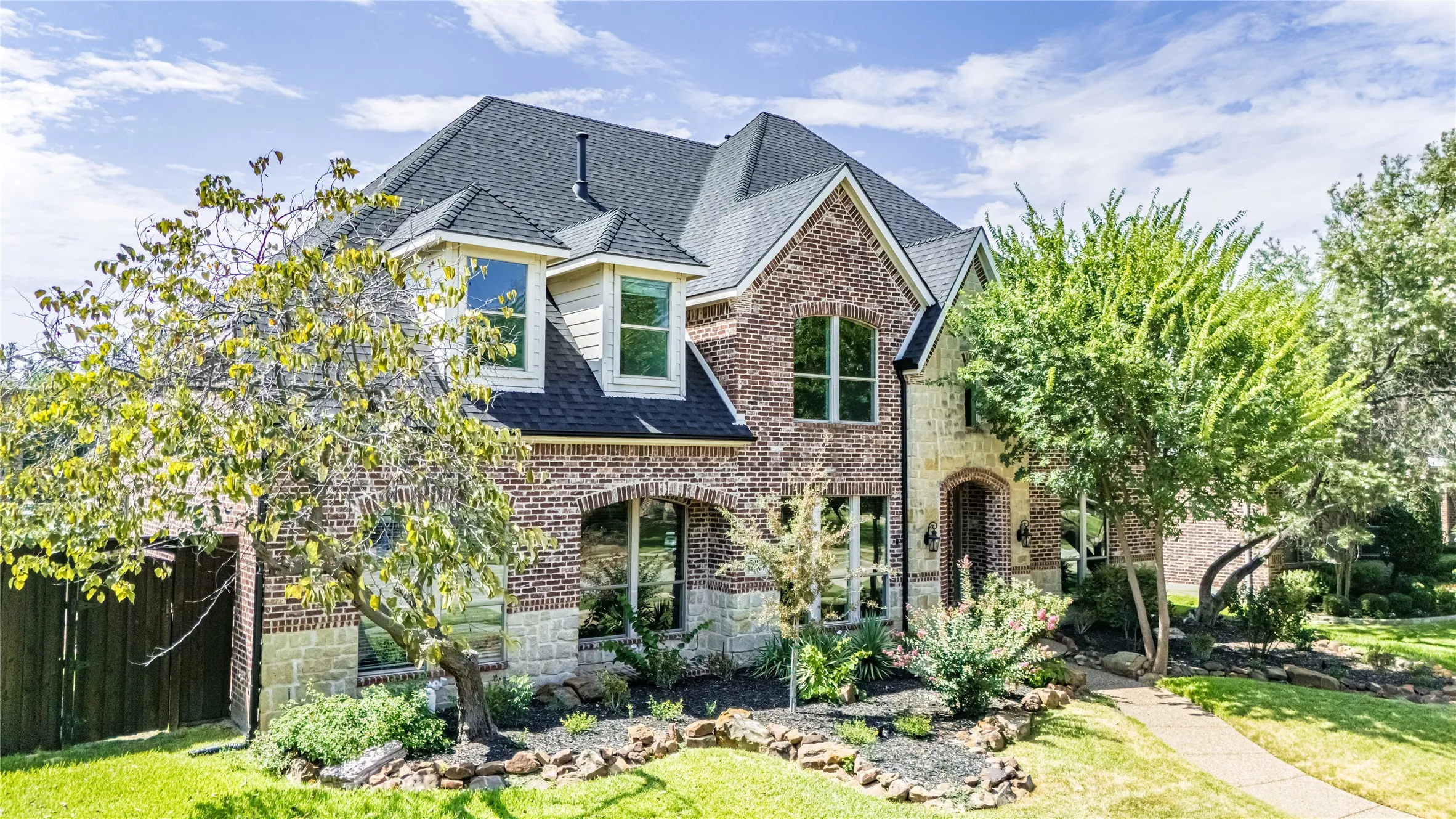Two-story residence featuring a brick and stone facade, arched windows, gabled rooflines, dark shingle roofing, and an inviting entry with a dark-stained door