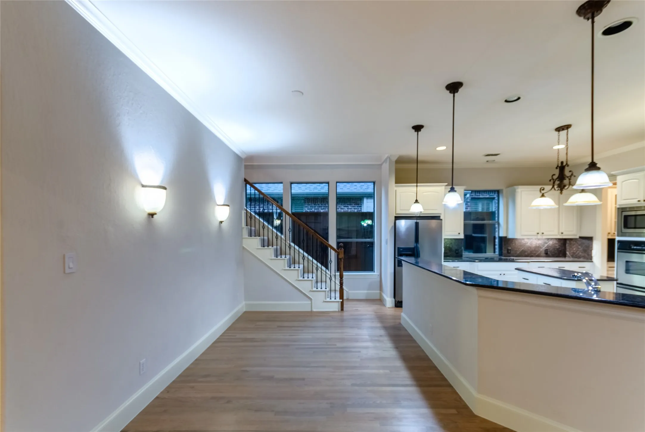 Open-concept living space featuring wood-finish flooring, a kitchen island with dark countertops, white cabinetry, stainless steel appliances, and a staircase with wrought iron balusters